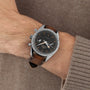 A close-up of a wrist wearing an OMEGA Speedmaster 57 with a brown leather strap, black dial, silver case, and Calibre 9906 movement over the sleeve of a brown knit sweater.