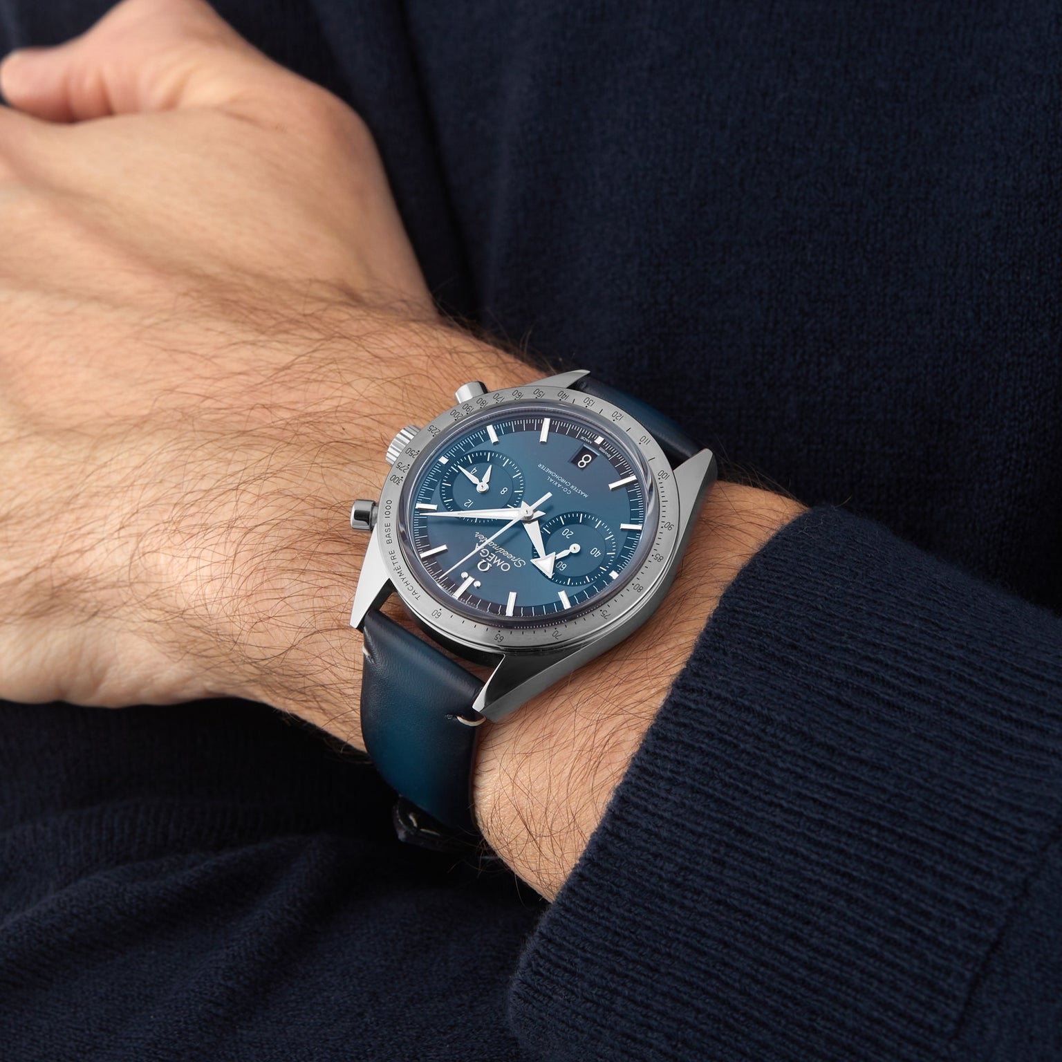 A person wearing an OMEGA Speedmaster 57 chronograph with a blue dial, leather strap, and a dark long-sleeve sweater.