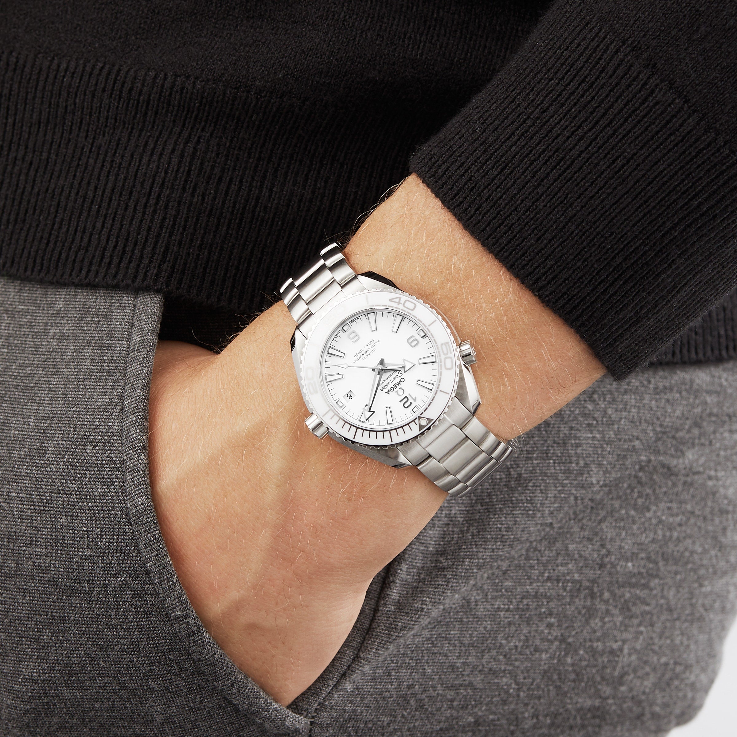 A person wearing an OMEGA Seamaster Planet Ocean 600M with a white dial and stainless steel case rests their hand in the pocket of grey pants, shown from the wrist down.