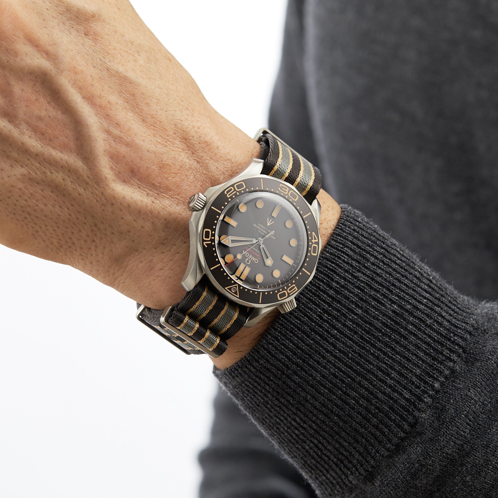 A person in a grey long-sleeve sweater wears an OMEGA Seamaster Diver 300M James Bond 007 Edition with a black dial and bezel, paired with a black and beige striped NATO strap.