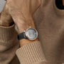 A person in a beige sweater showcases an OMEGA De Ville Prestige watch featuring a rose gold case, gray dial, and black leather strap on their left wrist.