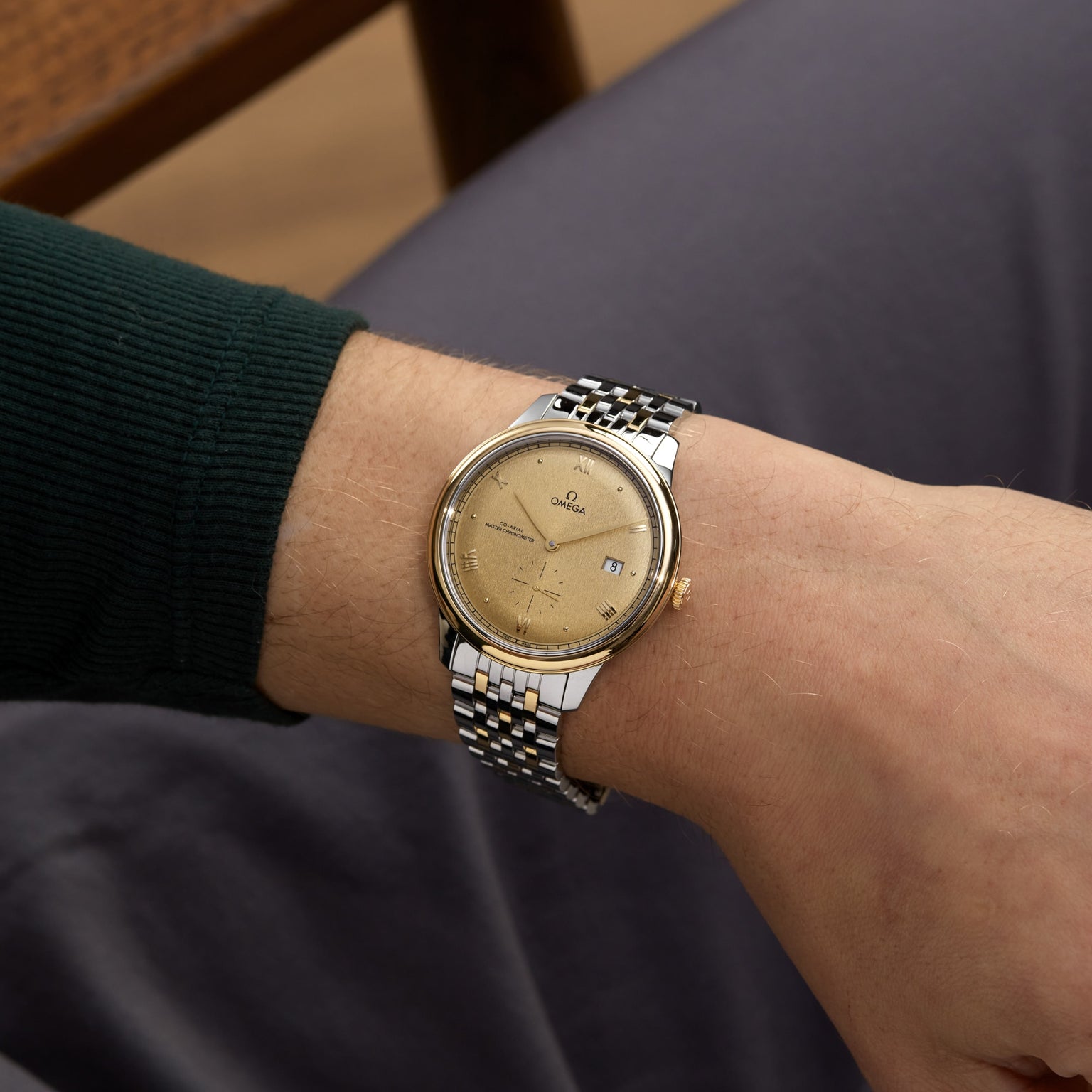 A person in a dark green long-sleeve shirt wears an OMEGA De Ville Prestige wristwatch featuring a Moonshine Gold dial and date display.