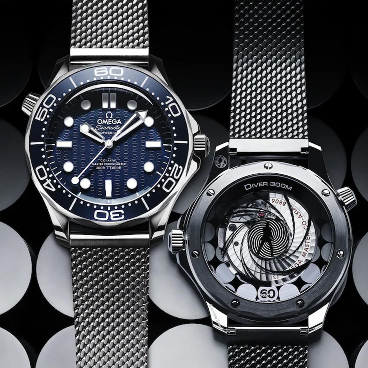 Two OMEGA Seamaster Diver 300M James Bond 007 60th Anniversary watches are shown: one with a blue patterned dial and bezel, the other displaying a transparent case back with Co-Axial movement. Both have sleek metal mesh bands.