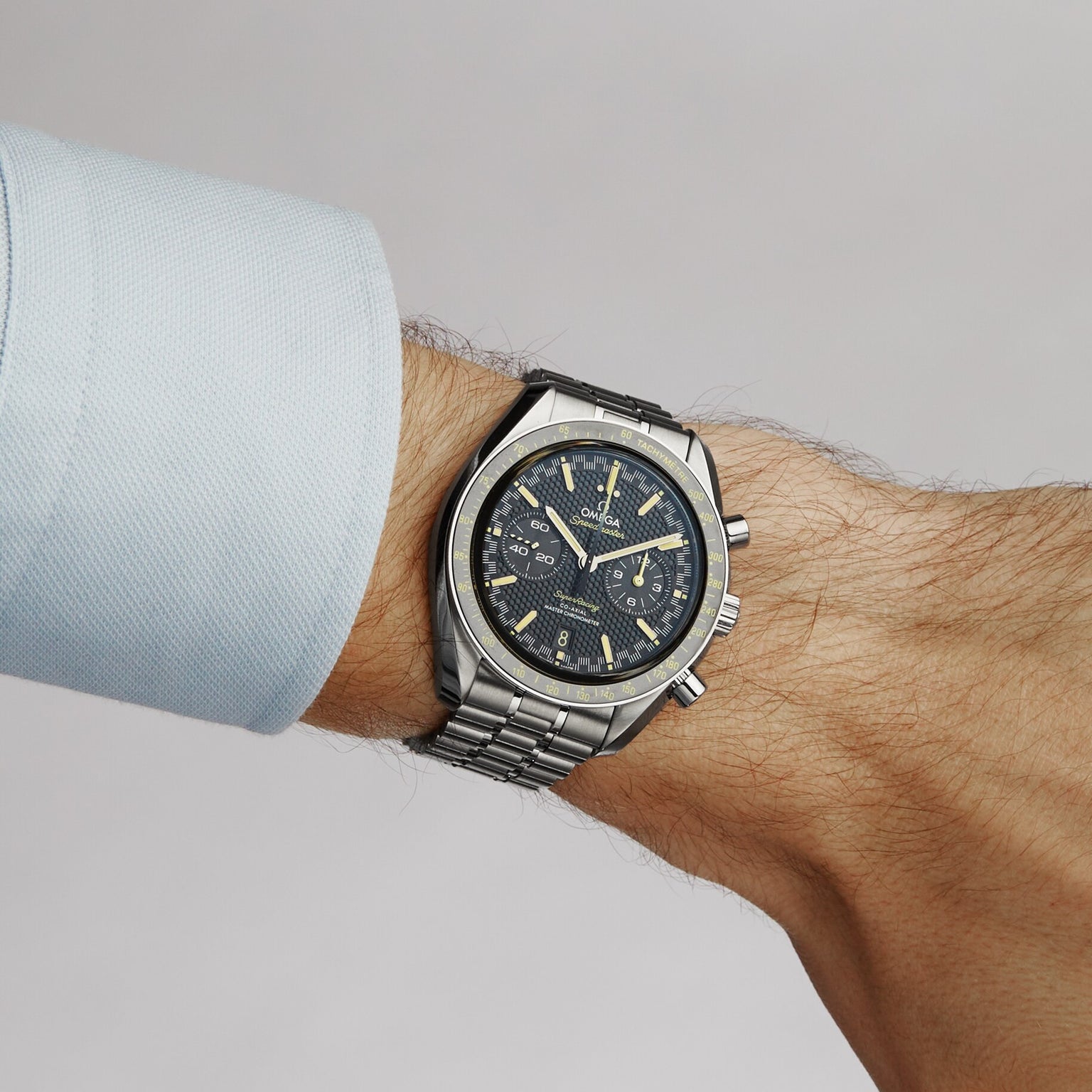 A person in a light blue shirt wears an OMEGA Speedmaster Super Racing chronograph with a black dial and tachymeter bezel on their left wrist.