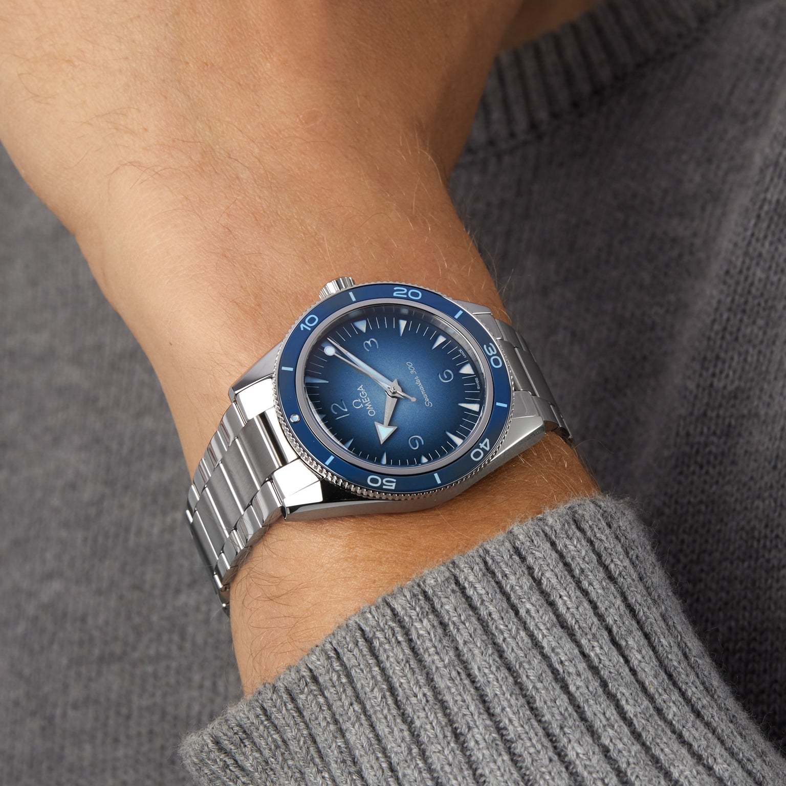 An individual wearing the OMEGA Seamaster 300 Summer Blue—with a blue dial and bezel, stainless steel silver bracelet, and Co-Axial Master Chronometer calibre 8912—on their left wrist, paired with a gray knit sweater.