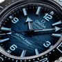 Close-up of the OMEGA Seamaster Planet Ocean 6000M watch face, featuring blue hands, numerals, and 6000m/20000ft on a textured blue background—an icon among saturation divers’ timepieces.