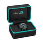An OMEGA Seamaster Planet Ocean 600M wristwatch with teal accents is displayed in an open black case with teal trim, featuring OMEGA and Emirates Team New Zealand logos inside the lid. Powered by the OMEGA 9900 calibre.