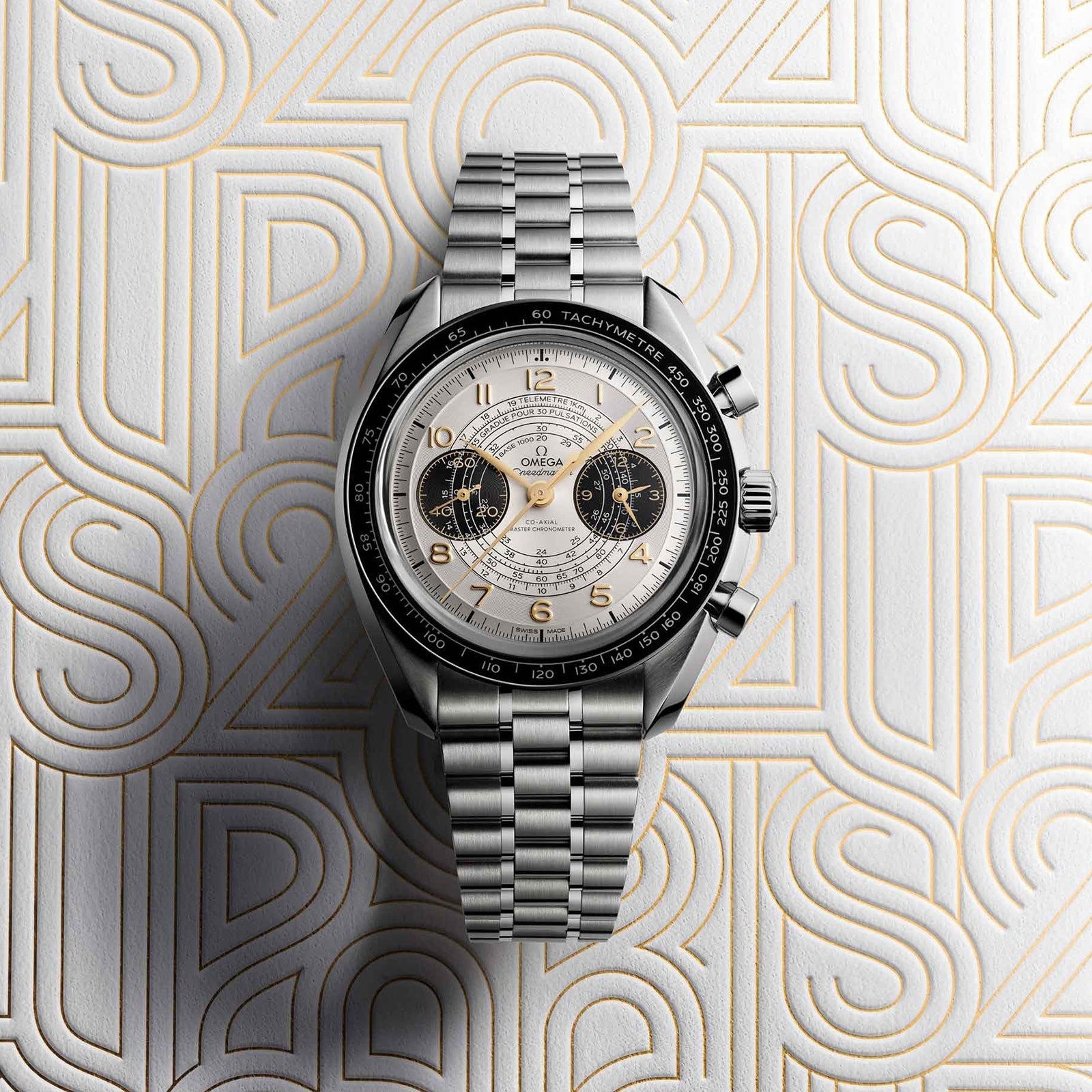 The OMEGA Speedmaster Chronoscope Paris 2024 Olympics wristwatch, featuring a silver case, white dial, and two black subdials, is displayed on a patterned background with raised gold and white lettering.
