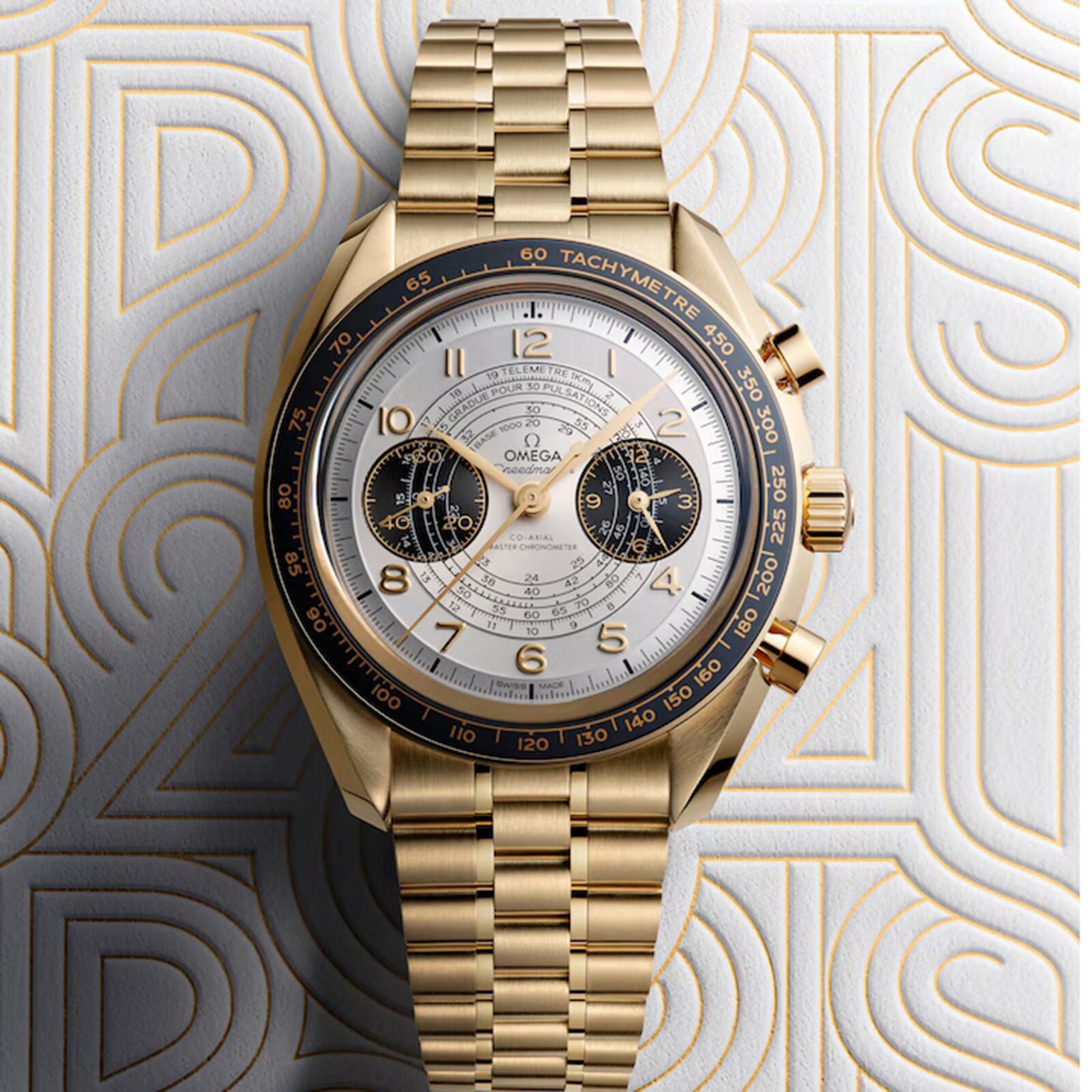 The OMEGA Speedmaster Chronoscope Paris 2024 Olympics features a gold case, white dial with two subdials, and a tachymeter bezel from the iconic Speedmaster collection, displayed on a patterned background.