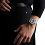 A person in black pants and a top rests their left hand near the back pocket, wearing an OMEGA Speedmaster 38 chronograph featuring a dark face with multiple subdials.