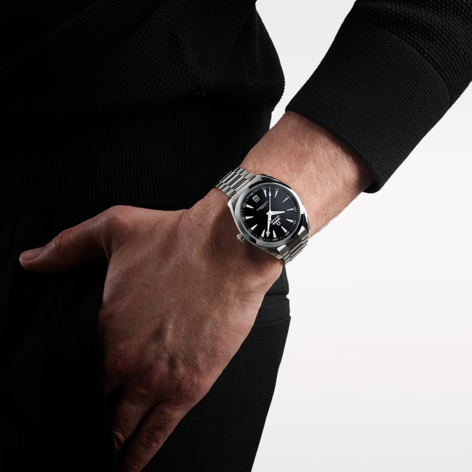 Wearing an OMEGA Seamaster Aqua Terra 150M with a black dial, this person rests their left hand on their hip and is dressed in a black long-sleeve shirt and pants.