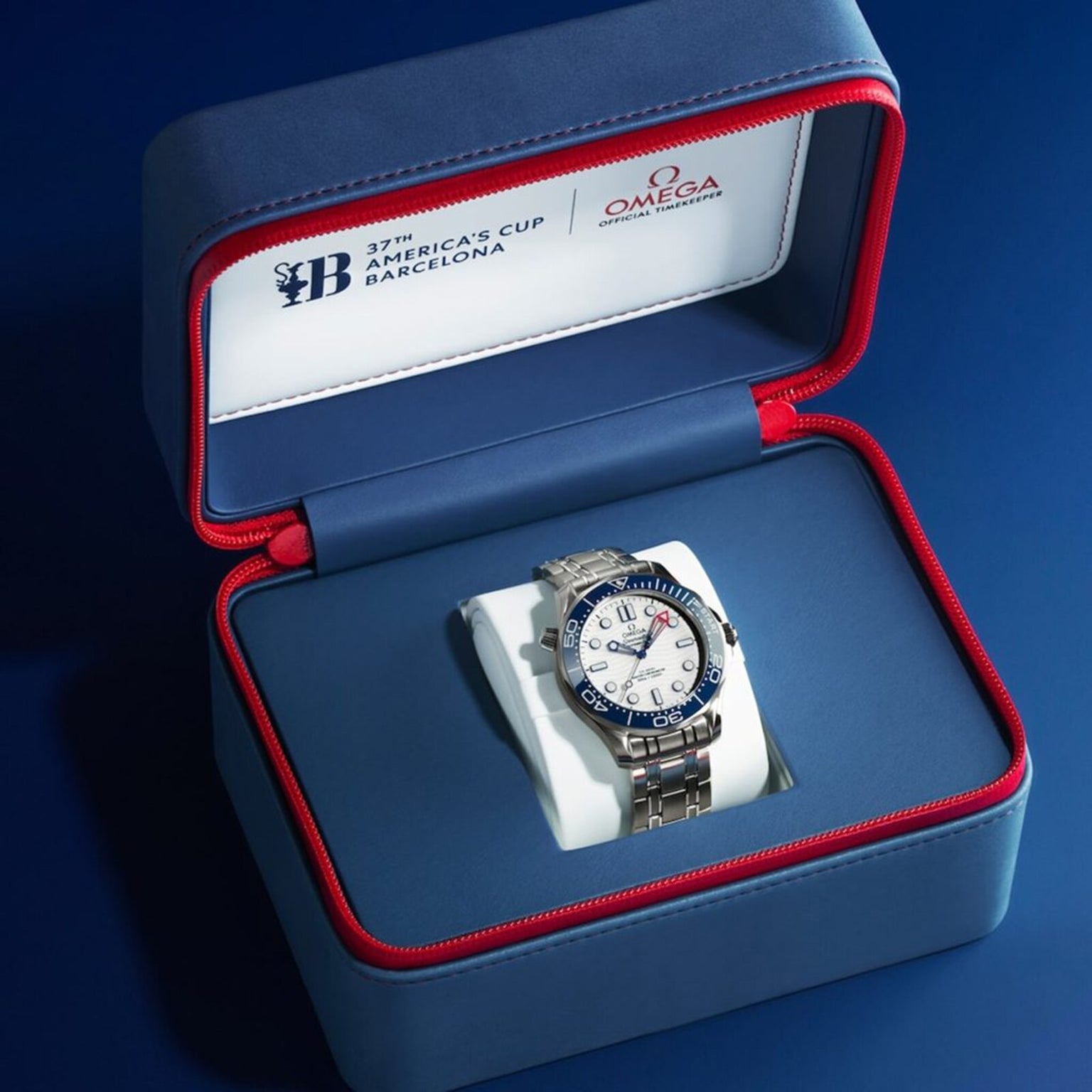 A silver OMEGA Seamaster Diver Americas Cup 300M with a white dial and blue bezel is shown in an open blue and red box marked 37th Americas Cup Barcelona.