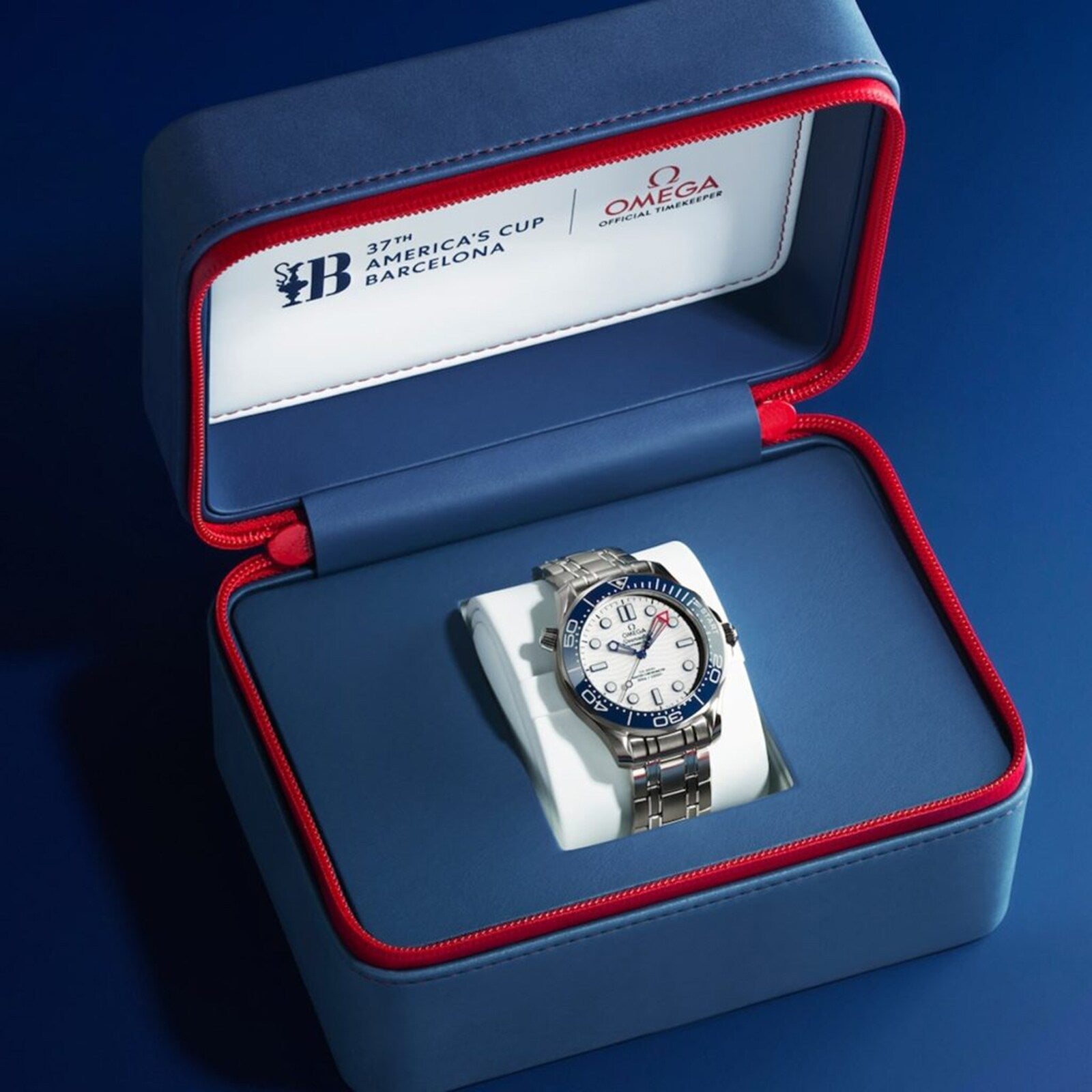 A silver OMEGA Seamaster Diver Americas Cup 300M with a white dial and blue bezel is shown in an open blue and red box marked 37th Americas Cup Barcelona.