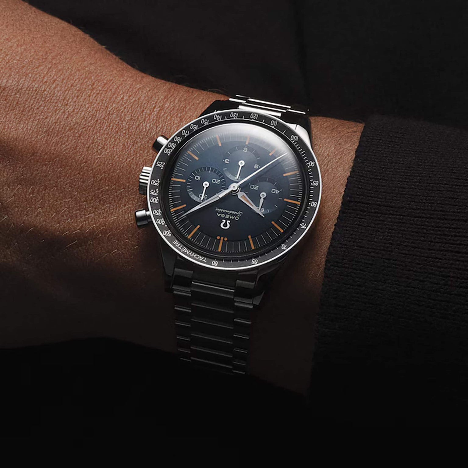 An OMEGA Speedmaster First OMEGA in Space stainless steel chronograph wristwatch with a black dial and orange accents is shown on a person’s wrist against a dark background.