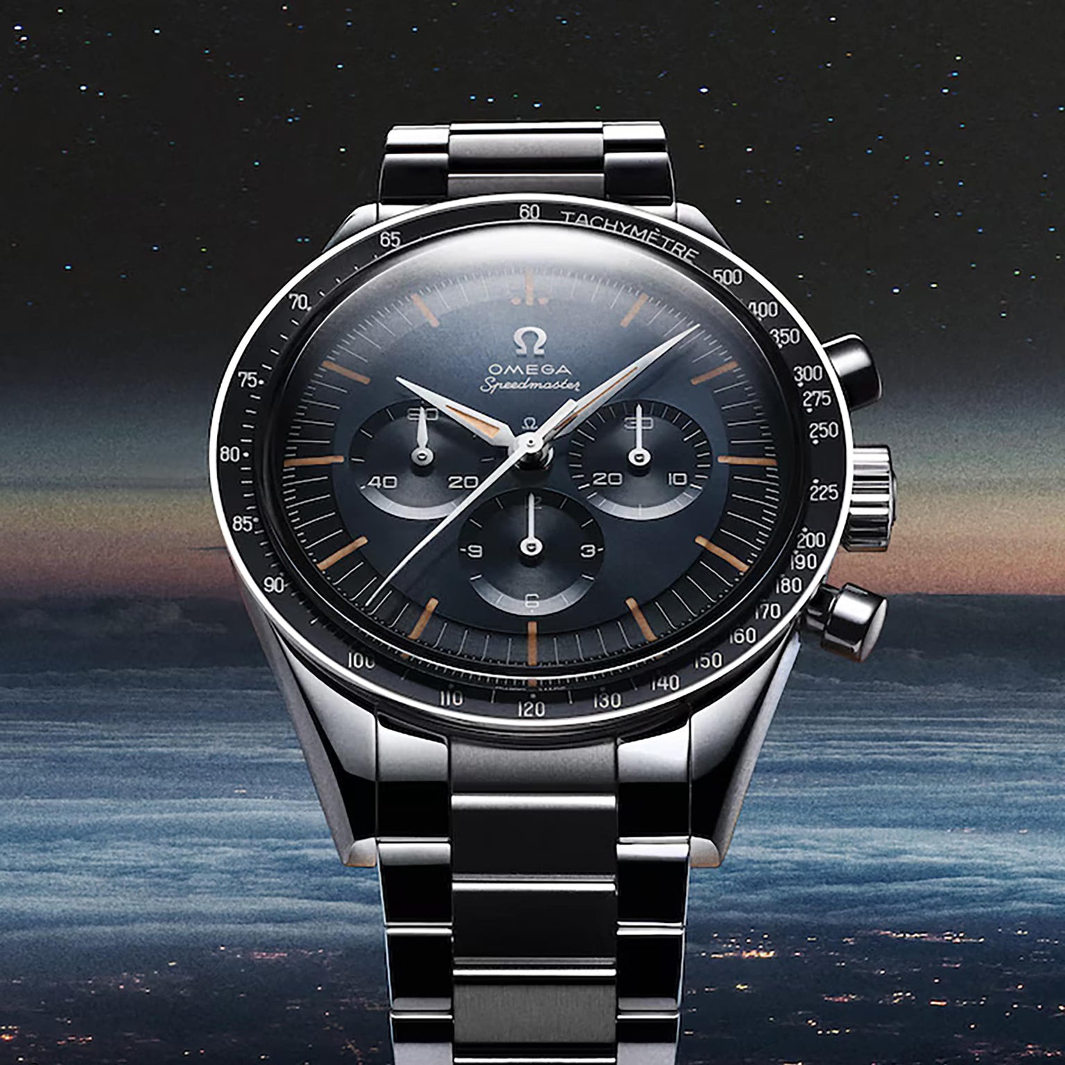 A stainless steel OMEGA Speedmaster First OMEGA in Space wristwatch with a black dial, three subdials, and tachymeter scale is photographed against a starry sky above city lights.