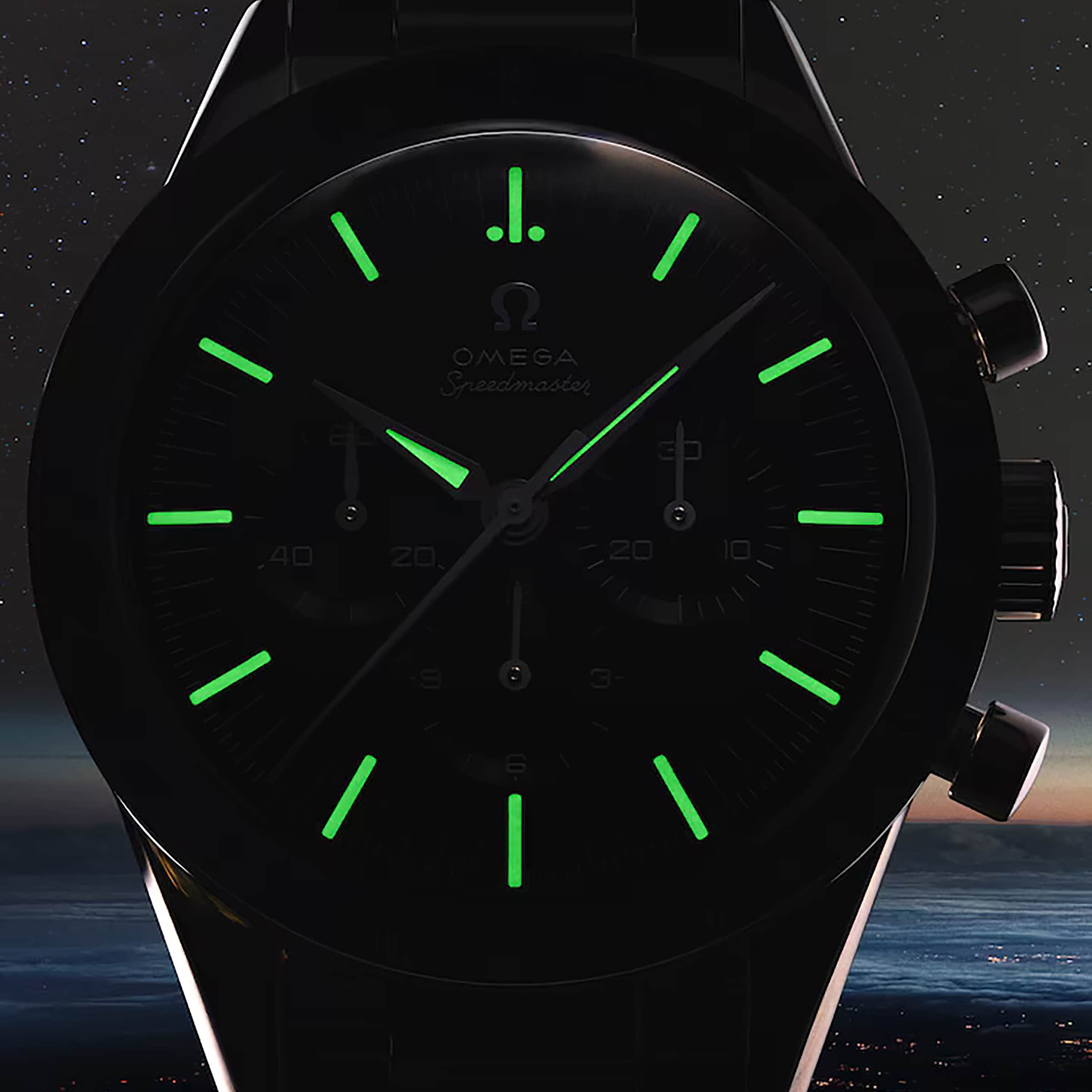 A close-up of an OMEGA Speedmaster First OMEGA in Space watch face in the dark, highlighting its glowing tachymeter scale and green hour markers and hands, set against a dim city lights backdrop with stars.