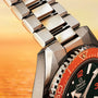 Close-up of a silver watch with a red and black bezel against an orange gradient background