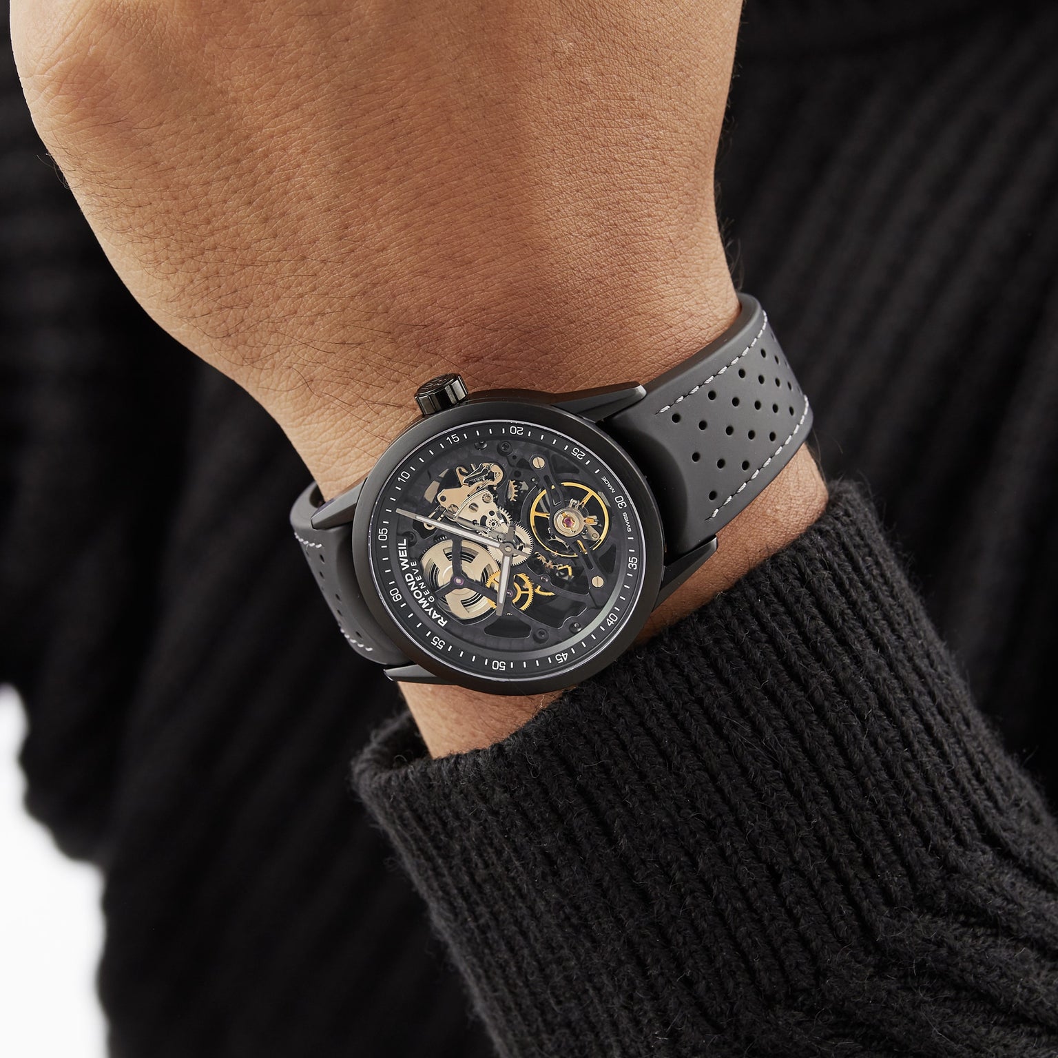A person wearing a Raymond Weil Freelancer Skeleton RW1212 Black Dial watch with a black perforated strap on their left wrist, dressed in a black ribbed sweater.