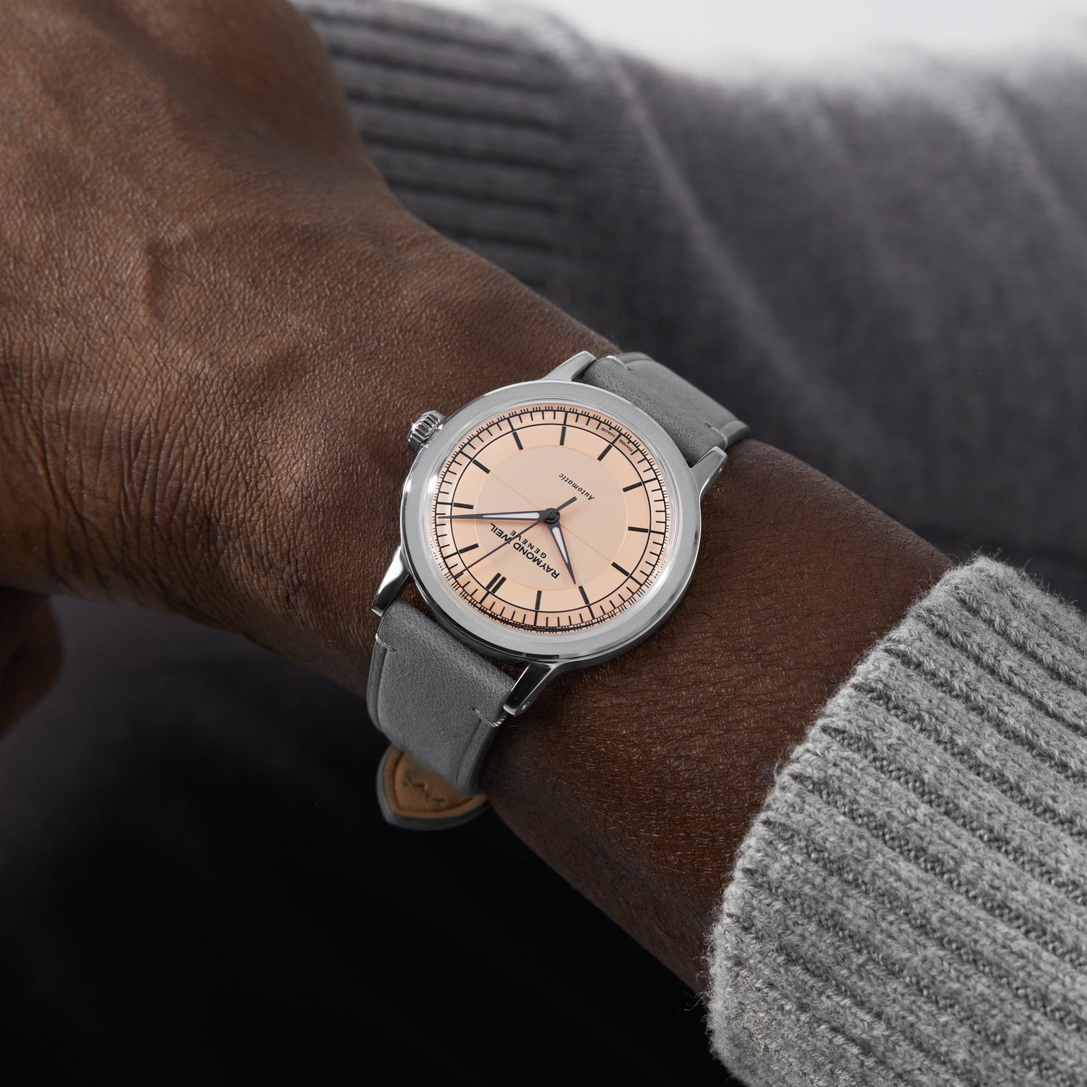 A person wearing a Raymond Weil Millesime Automatic Salmon Sector Dial Swiss-made silver wristwatch with a grey leather strap, dressed in a grey sweater.
