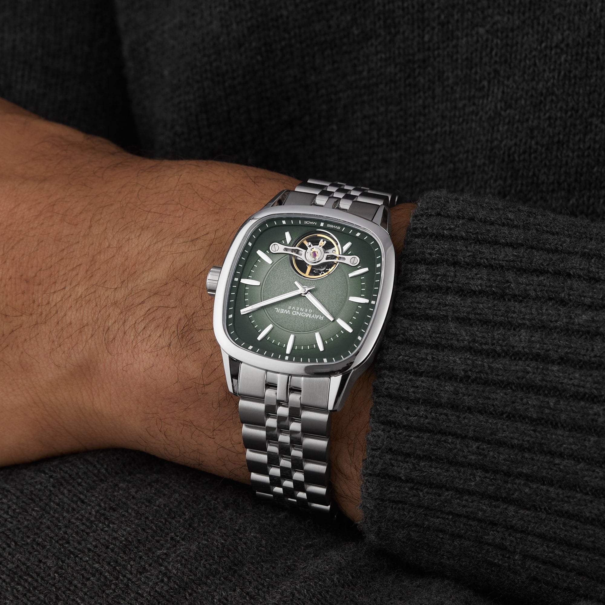 A person wearing a Raymond Weil Freelancer Calibre RW1212 Automatic with a green dial and metallic link band, dressed in a dark knit sweater.