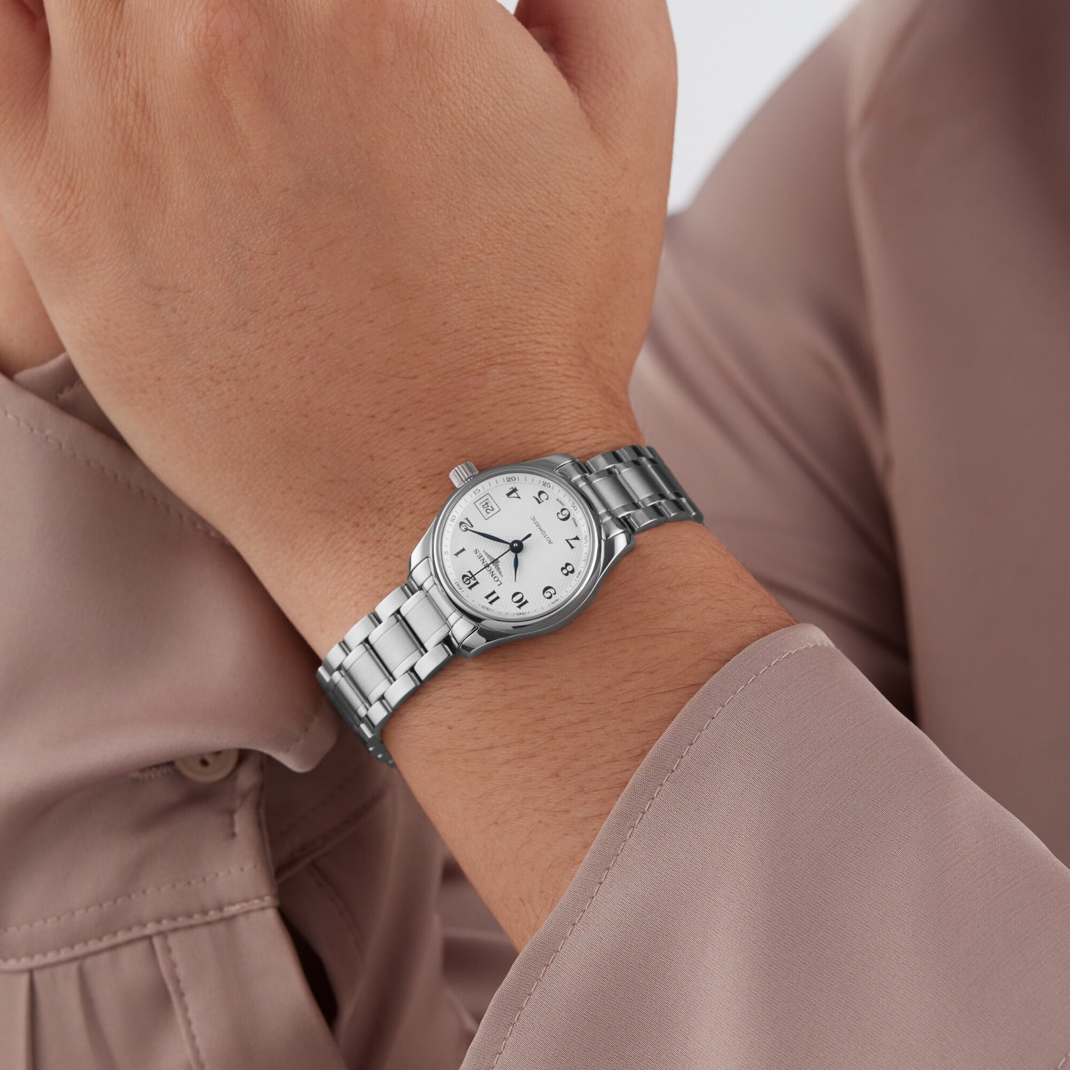 A person wearing a Longines Master Collection wristwatch with a white face and stainless steel bracelet, paired with a light beige long-sleeve shirt.