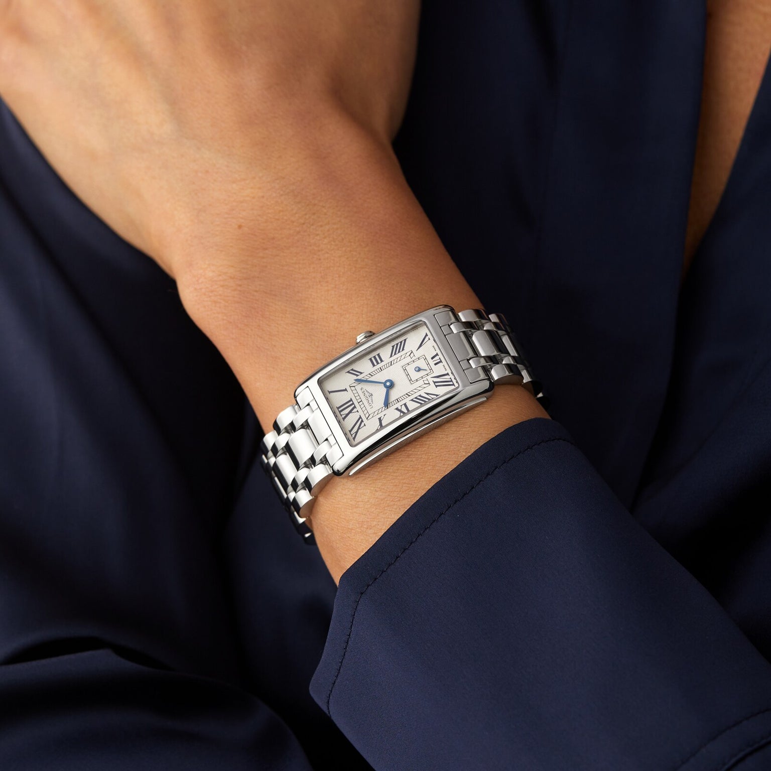 A person dressed in a dark blue long-sleeve garment wears the Longines DolceVita, featuring an elegant rectangular stainless steel case and a dial with Roman numerals.