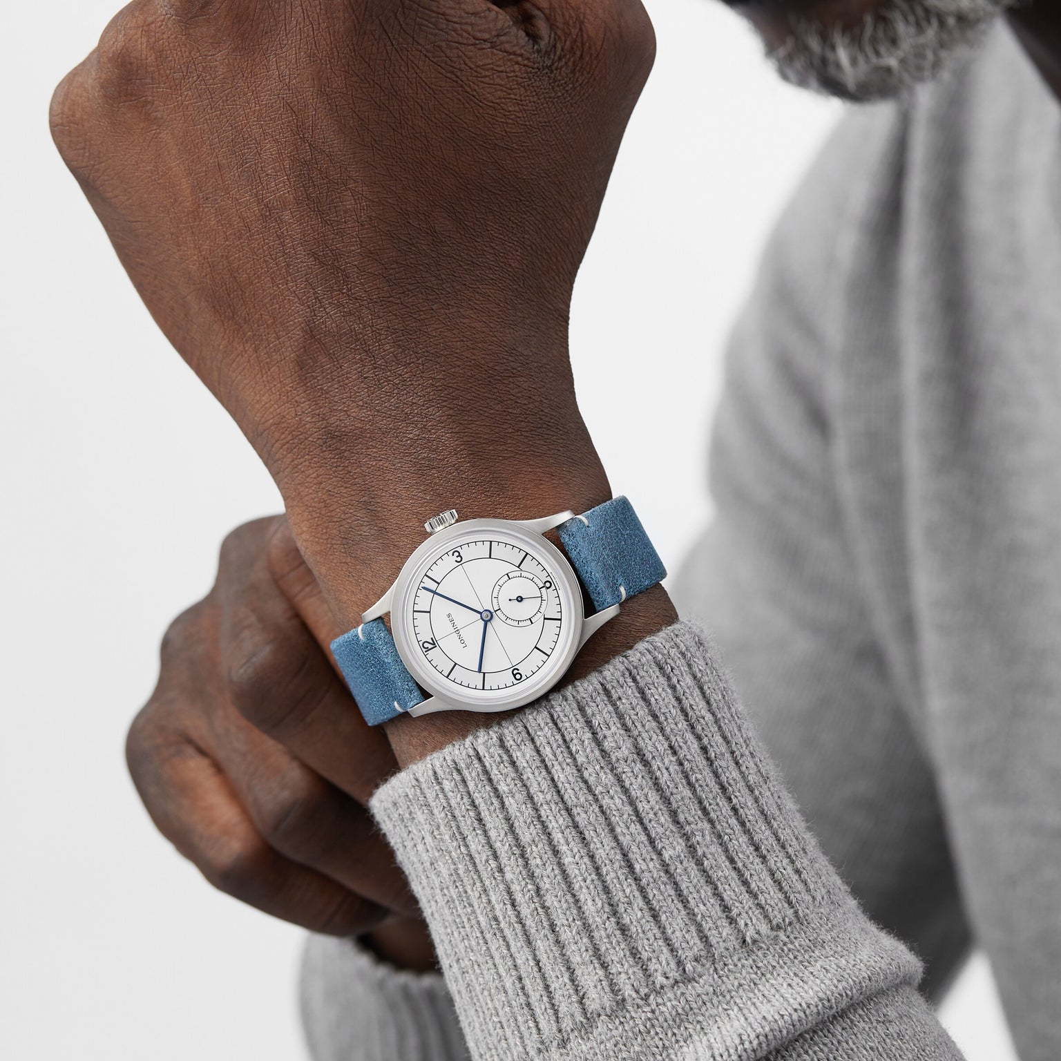 A person wearing a Longines Sector Dial silver wristwatch with a white face and blue strap, featuring a stainless steel case, dressed in a light gray sweater.