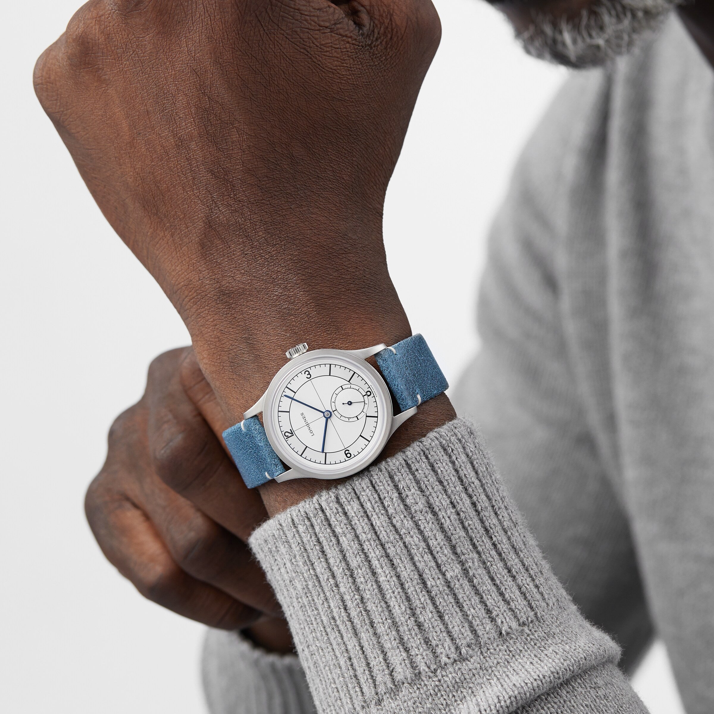 A person wearing a Longines Sector Dial silver wristwatch with a white face and blue strap, featuring a stainless steel case, dressed in a light gray sweater.