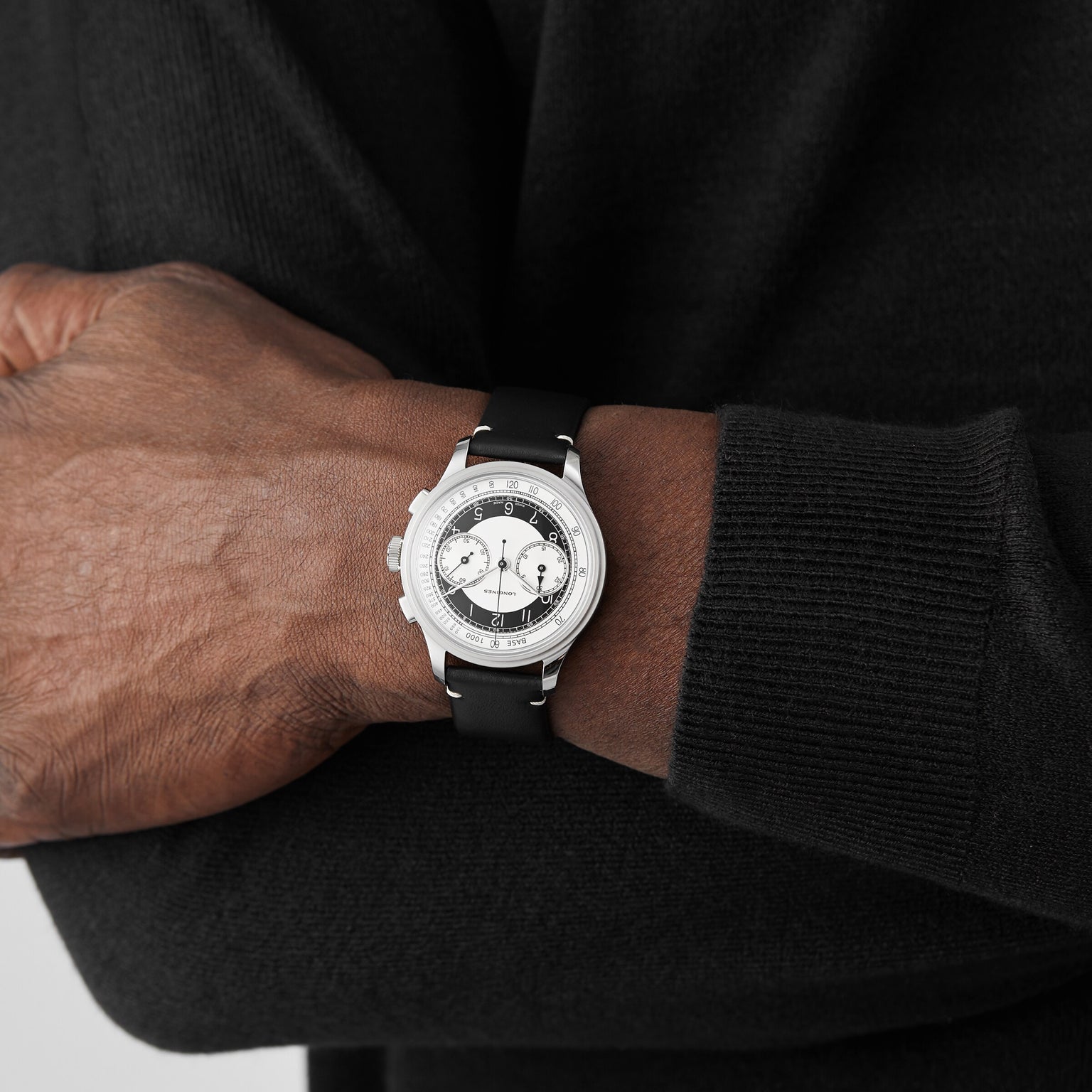 A person in a black sweater crosses their arms, showcasing the Longines Tuxedo wristwatch from the Heritage Classic collection, which features a stainless steel case and a white and black dial.