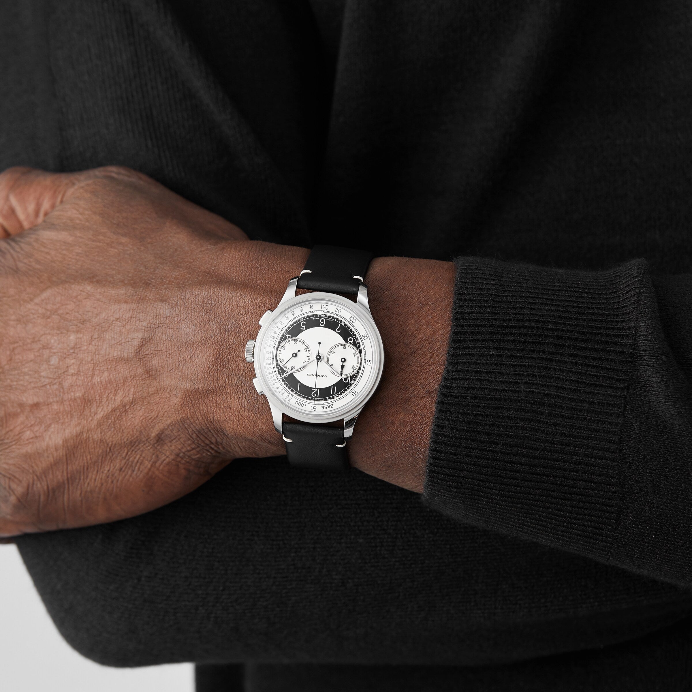 A person in a black sweater crosses their arms, showcasing the Longines Tuxedo wristwatch from the Heritage Classic collection, which features a stainless steel case and a white and black dial.