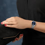 Wearing the Longines Master Collection silver watch with a blue face, a person holds a black clutch purse against a blurred blue background.