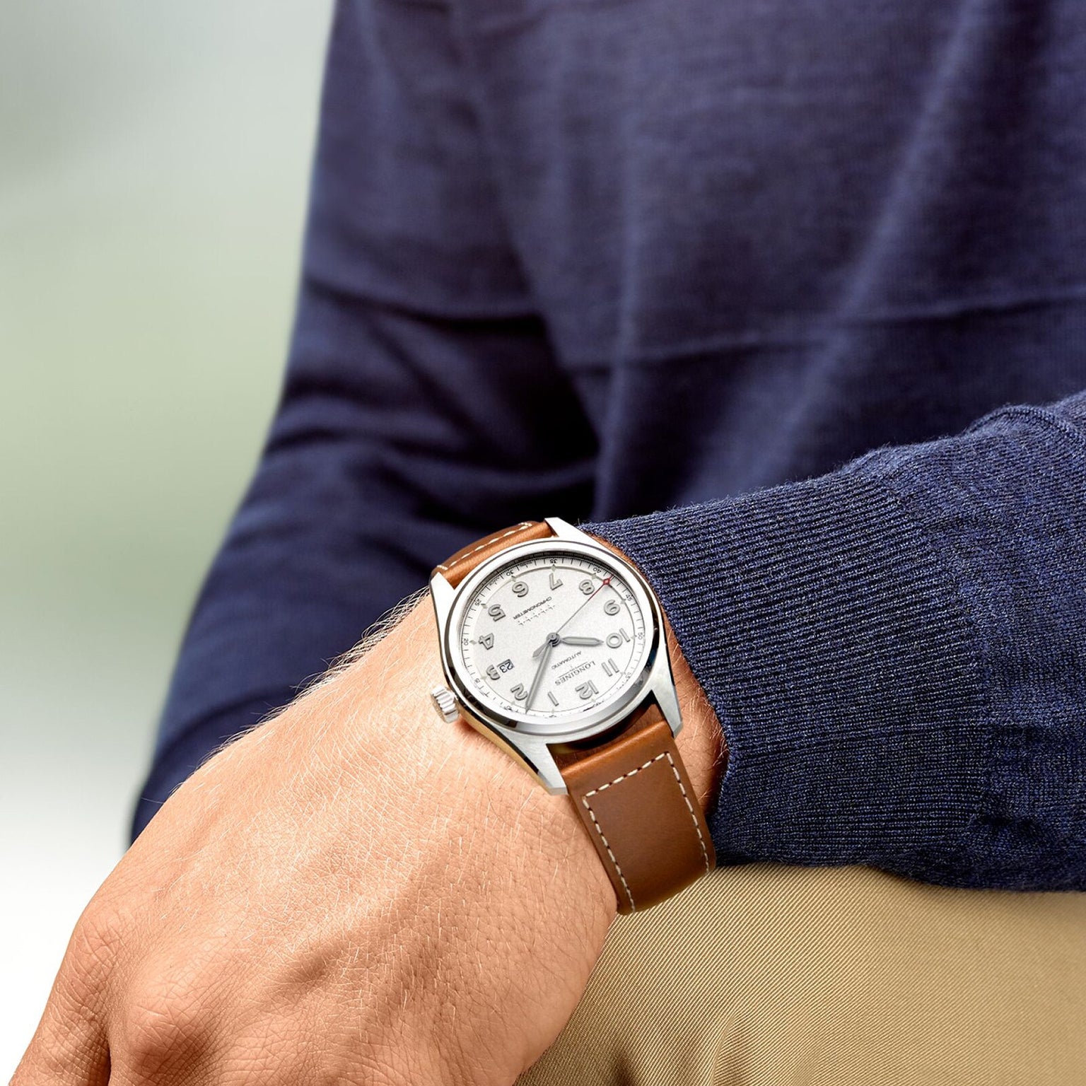 A person in a blue sweater and beige pants wears the Longines Spirit, an analog wristwatch with a white face and brown leather strap, on their left wrist.
