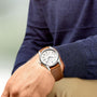 A person in a blue sweater and beige pants wears the Longines Spirit, an analog wristwatch with a white face and brown leather strap, on their left wrist.