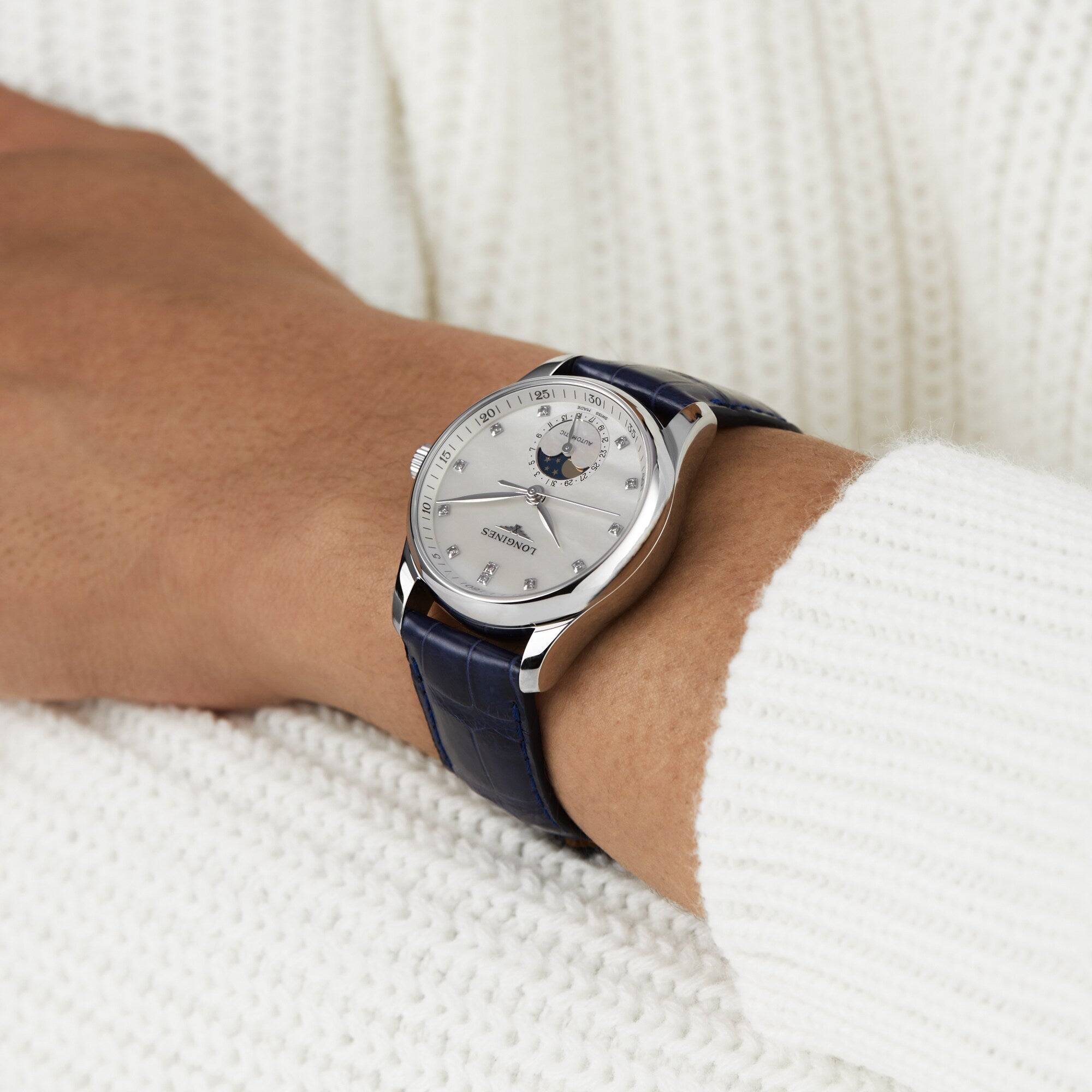 A person wearing the Longines Master Collection Moonphase, a silver wristwatch with a blue leather strap and white face featuring diamond indexes, displayed on their left wrist over a white knit sweater.