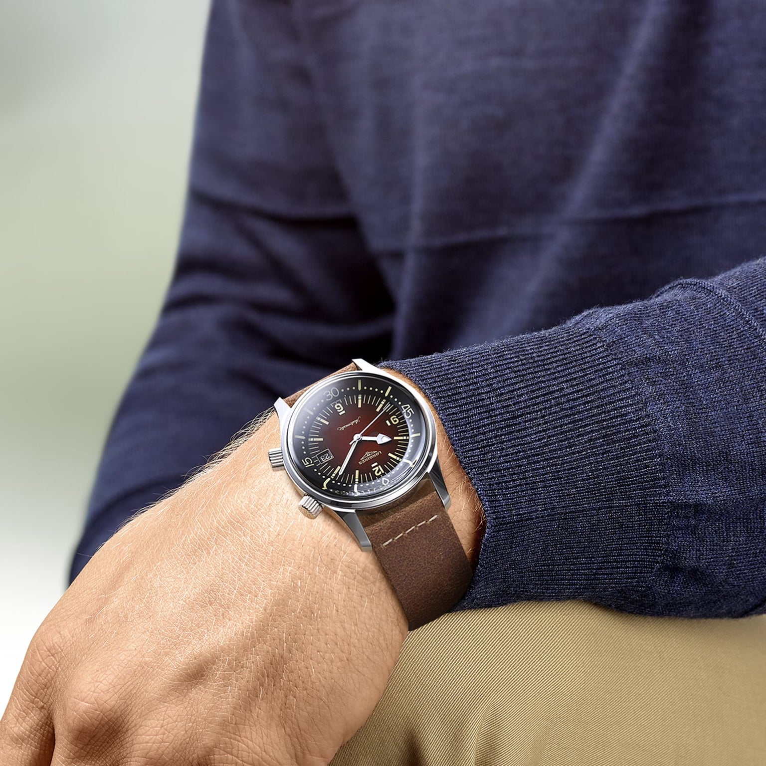 Someone in a dark blue sweater and beige pants displays a Longines Legend Diver watch with a brown leather strap and black dial on their left wrist.
