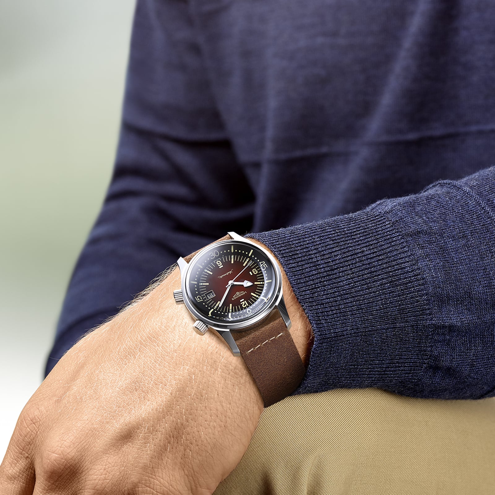 Someone in a dark blue sweater and beige pants displays a Longines Legend Diver watch with a brown leather strap and black dial on their left wrist.
