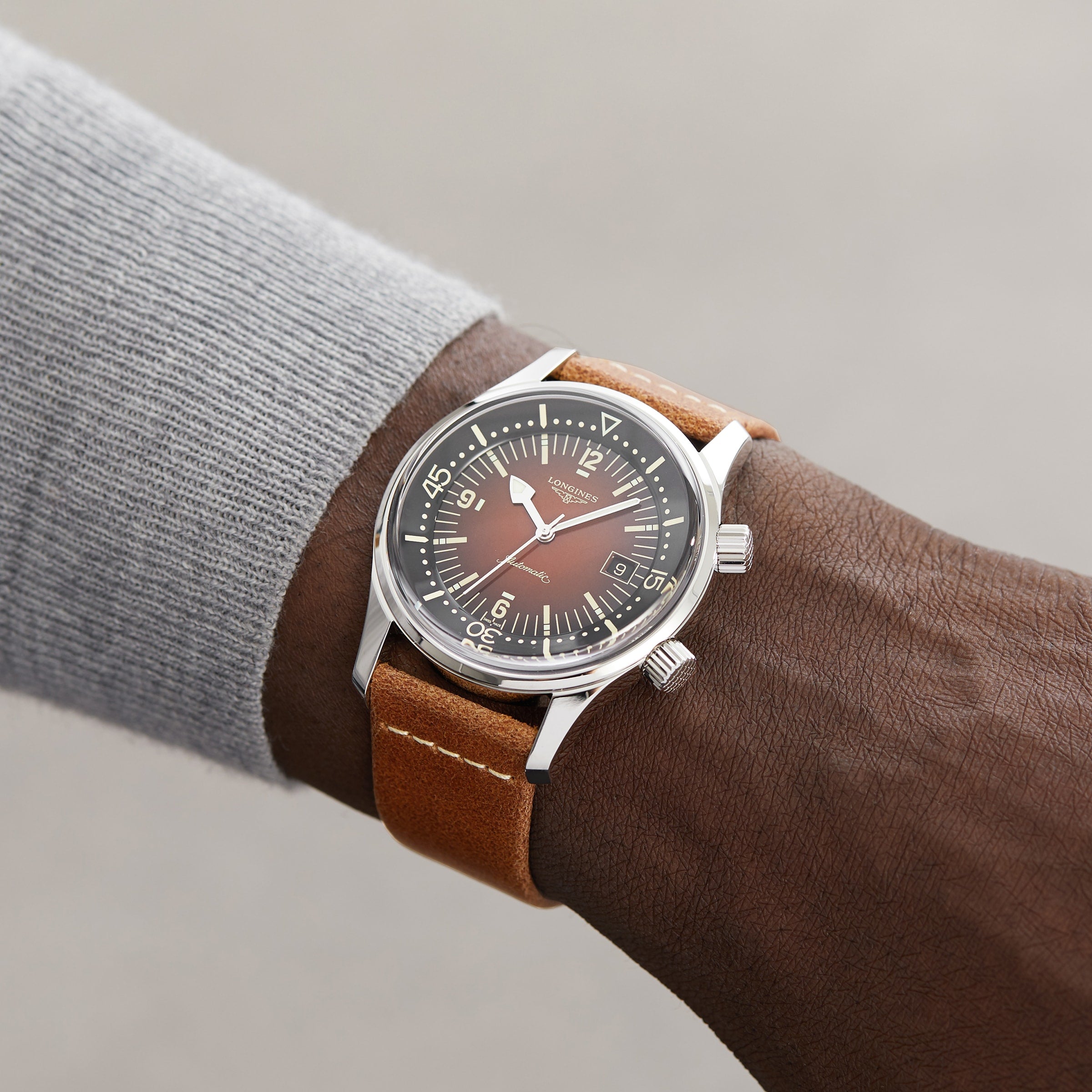 A person wears a Longines Legend Diver watch with a brown leather strap and black dial, showing 10:07. The watch pairs perfectly with their light gray long-sleeve shirt.