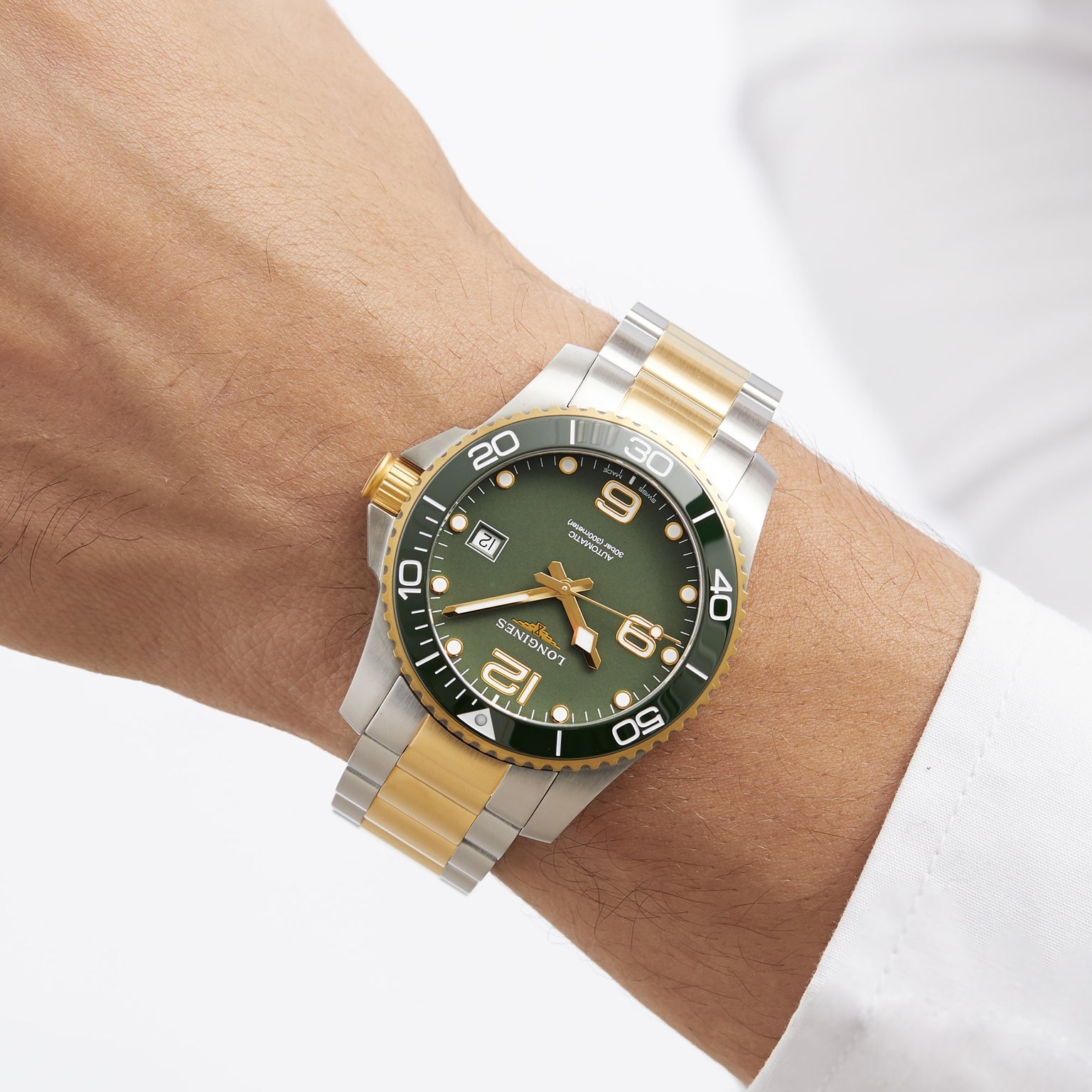 A wrist wearing a Longines HydroConquest two-tone metal diving watch with a green dial, gold accents, and bold white numerals is shown. The person is dressed in a white shirt.