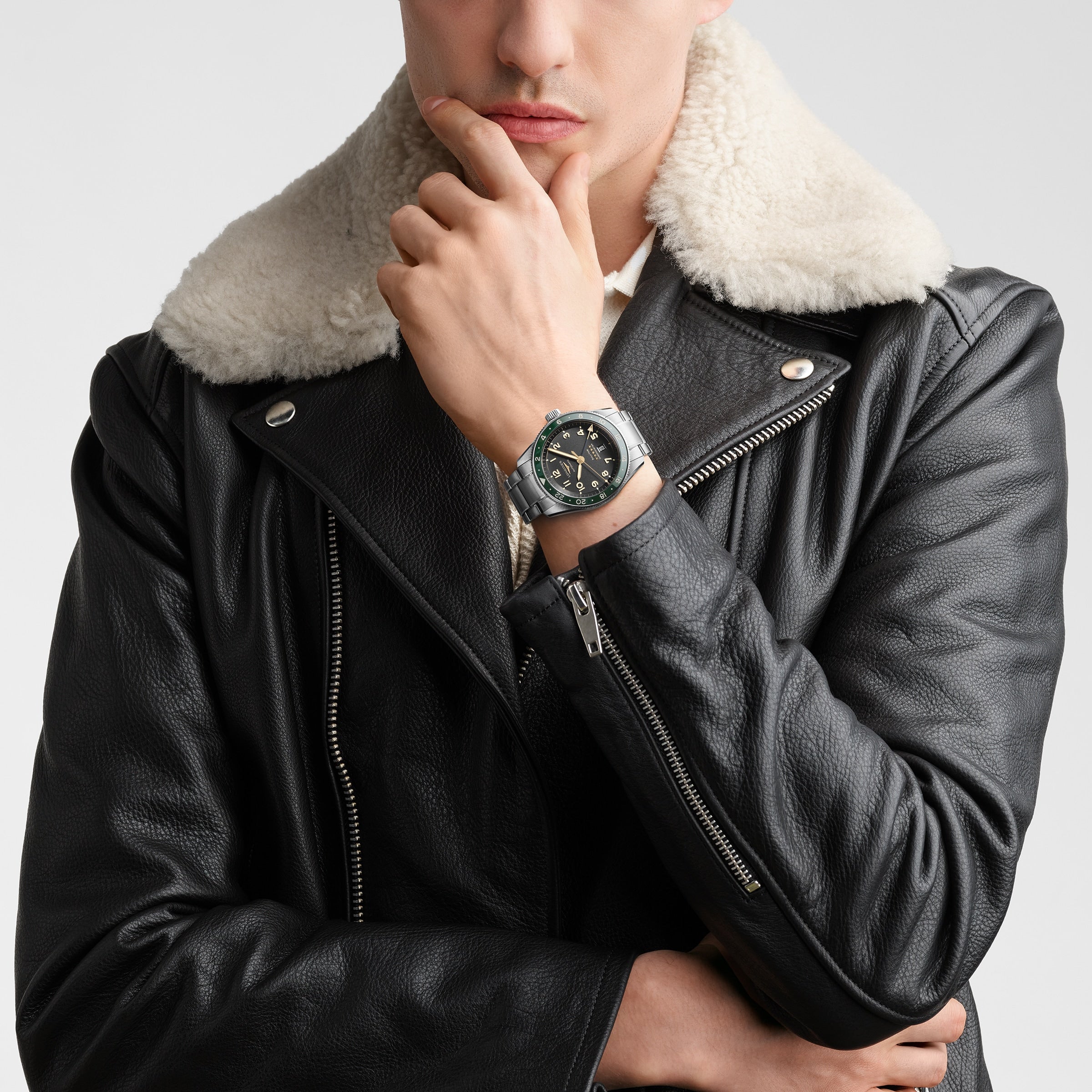 A person in a black leather jacket with a shearling collar poses with their hand on their chin, showcasing the Longines Spirit Zulu Time watch.