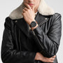 A person in a black leather jacket with a shearling collar, hand on chin, wears the Longines Spirit Zulu Time wristwatch featuring a stainless steel case, black dial, and brown strap.