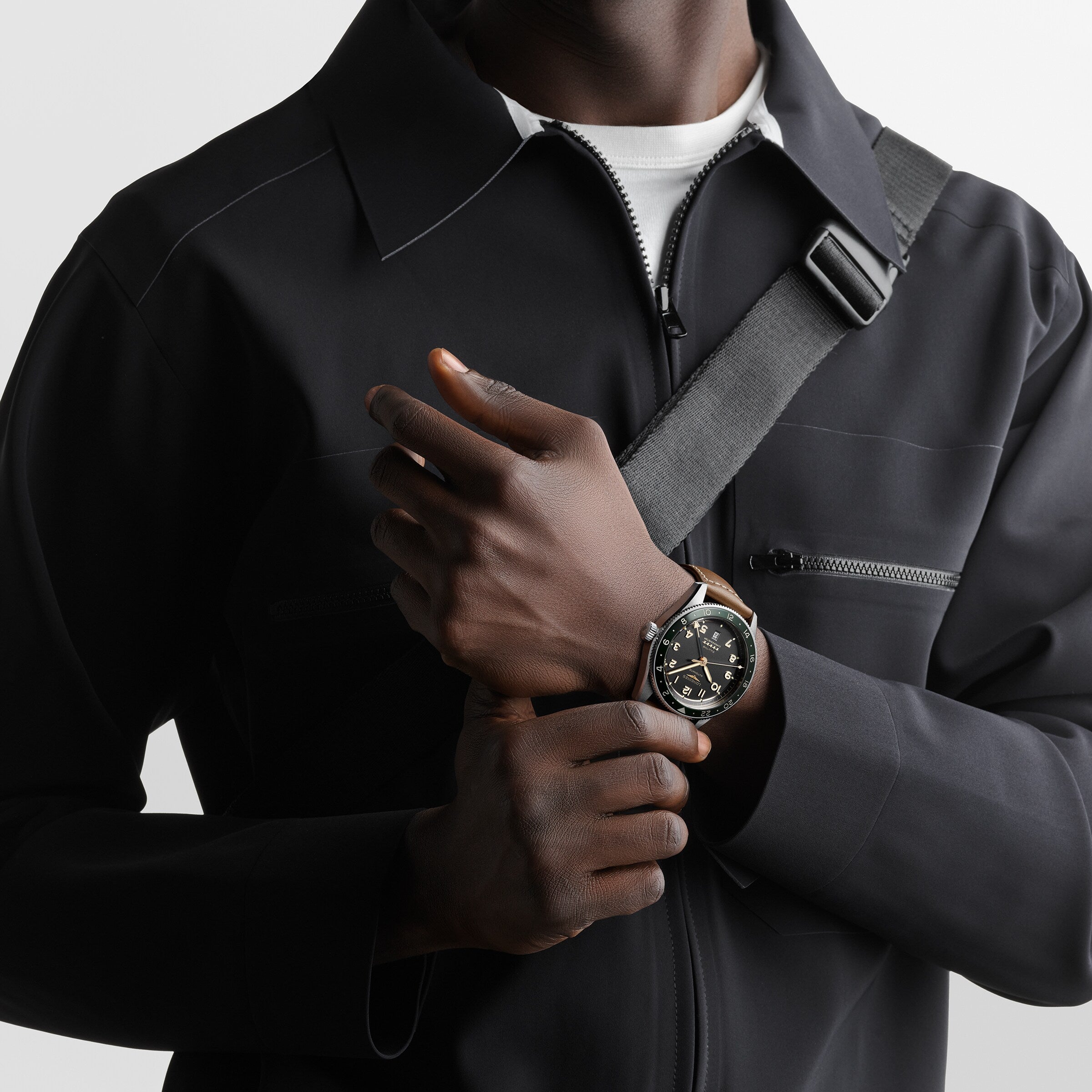 A person in a black zip-up jacket with a crossbody strap holds their wrist, showcasing the Longines Spirit Zulu Time watch, featuring a black dial and stainless steel case.