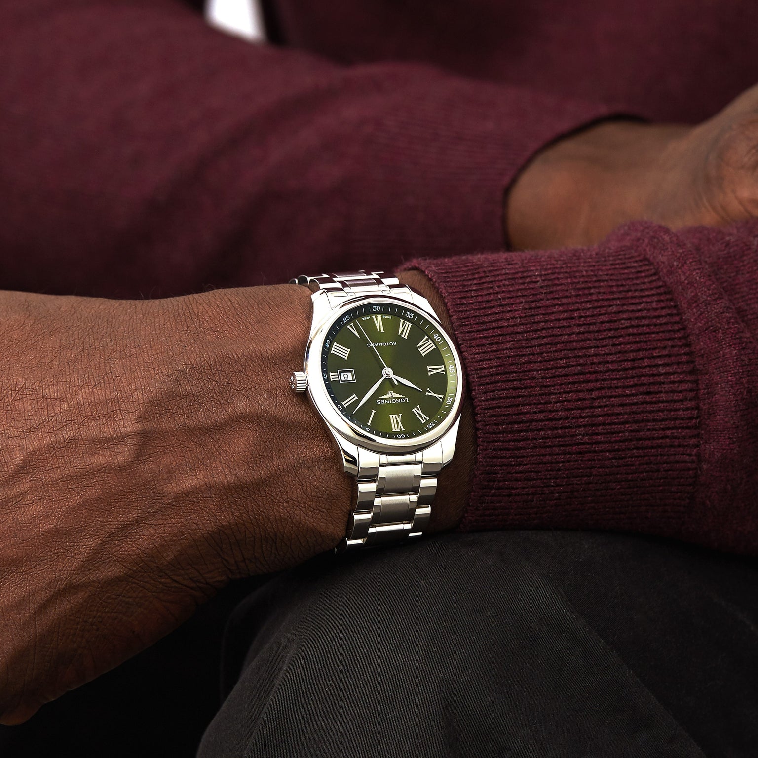 A person in a burgundy long-sleeve shirt rests their arm on their lap, proudly wearing the Longines Master Collection silver wristwatch with a green dial and Roman numerals.