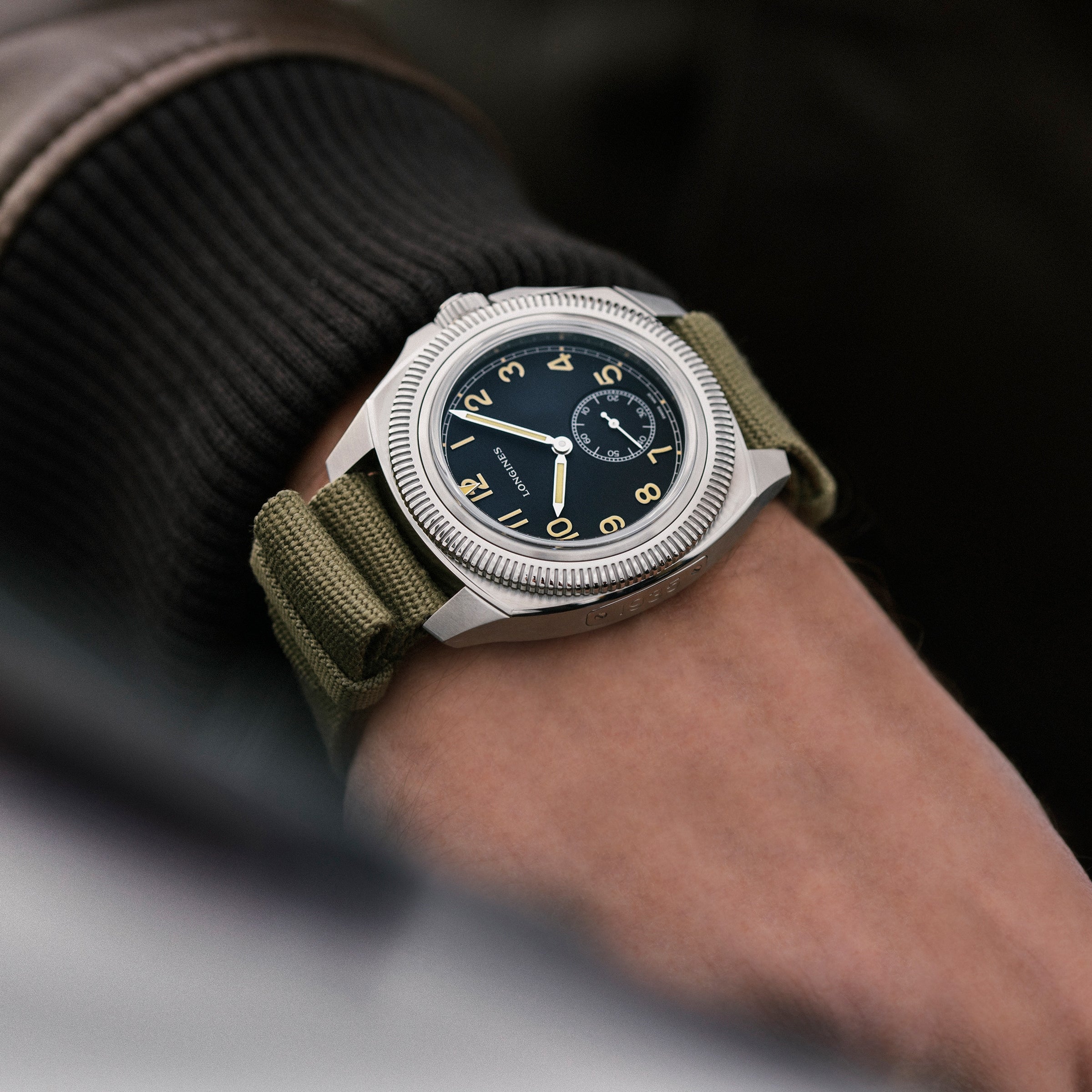A wrist sporting the Longines Pilot Majetek, an aviation-inspired watch with a green fabric strap, black dial, and yellow numerals, peeks from beneath a dark ribbed sleeve.