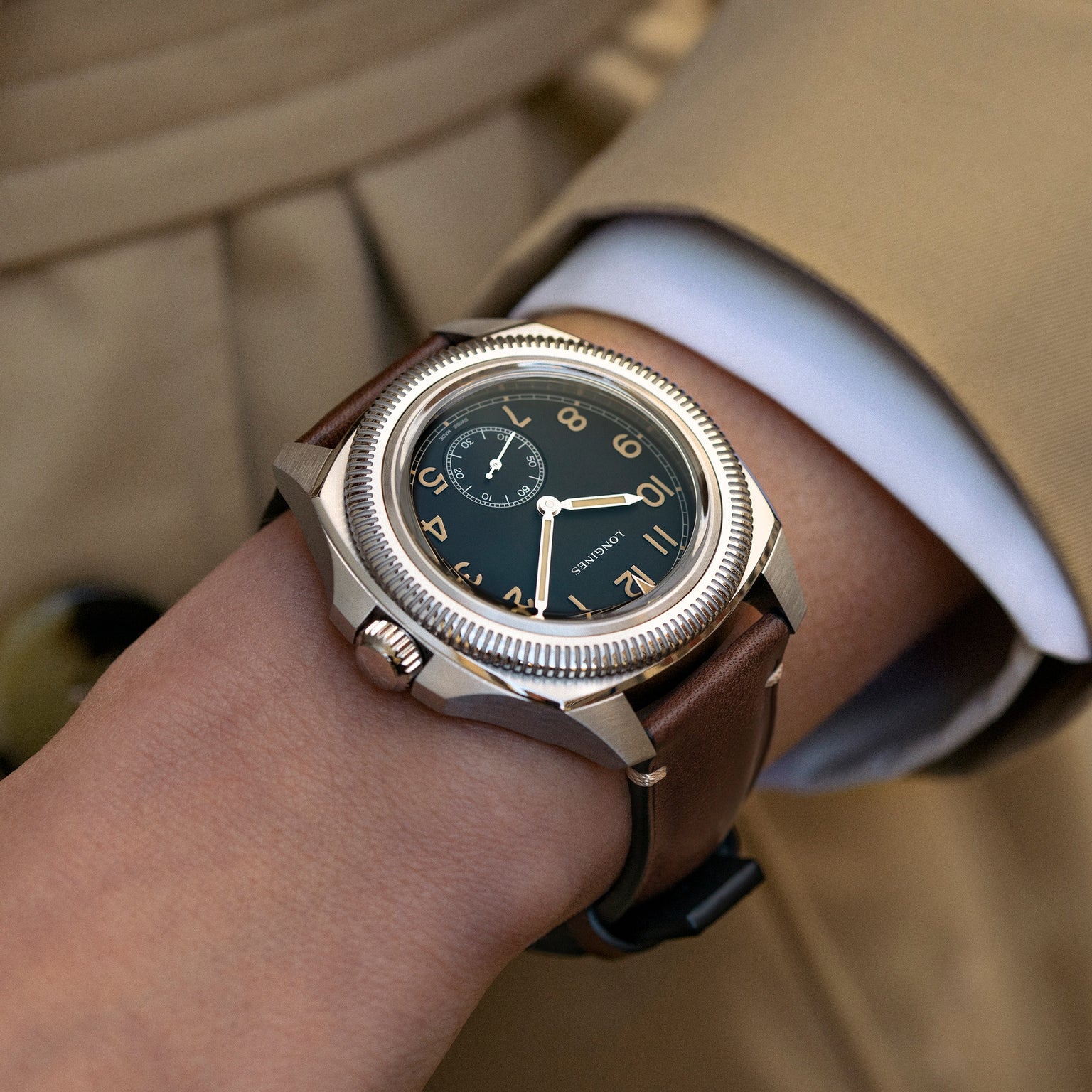 A close-up of a person wearing the Longines Pilot Majetek, featuring a brown leather strap, black dial, gold numerals, and small seconds subdial—reflecting aviation heritage against a tan sleeve.