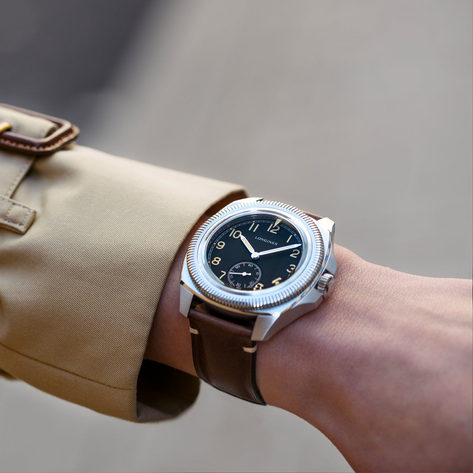 A person in a beige coat showcases the Longines Pilot Majetek, featuring a black dial, gold numerals, and a brown leather strap—an elegant nod to classic aviation heritage.