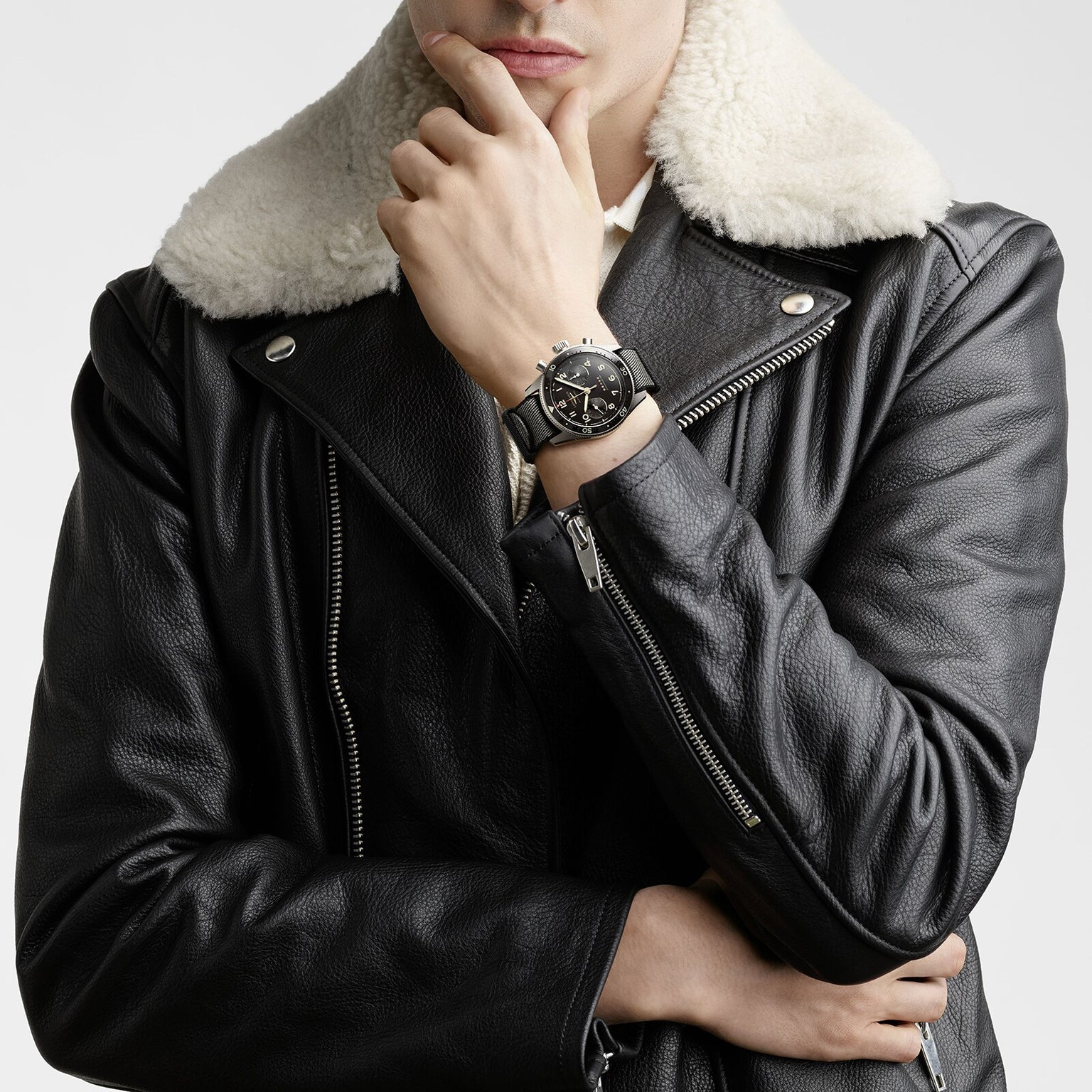 A person in a black leather jacket with a white shearling collar, arms crossed and hand on chin, showcases the Longines Spirit Flyback Titanium pilot watch.