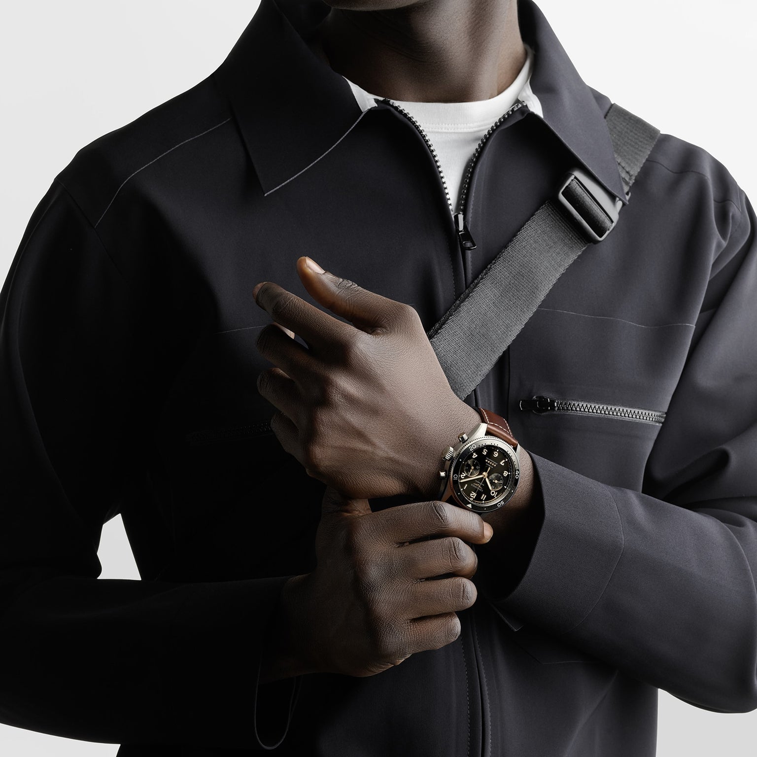 A person in a black zip-up jacket with a crossbody strap showcases the Longines Spirit Flyback watch, featuring a brown strap and round black dial with COSC-certified chronograph movement.