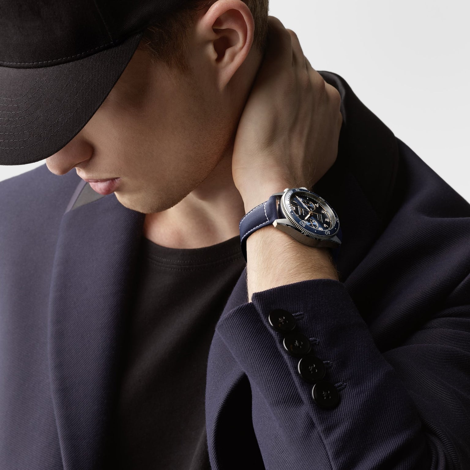 A man in a navy blazer and black cap touches his neck, wearing the Longines Spirit Flyback—a silver wristwatch with a blue strap.