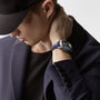 A man in a navy blazer and black cap touches his neck, wearing the Longines Spirit Flyback—a silver wristwatch with a blue strap.