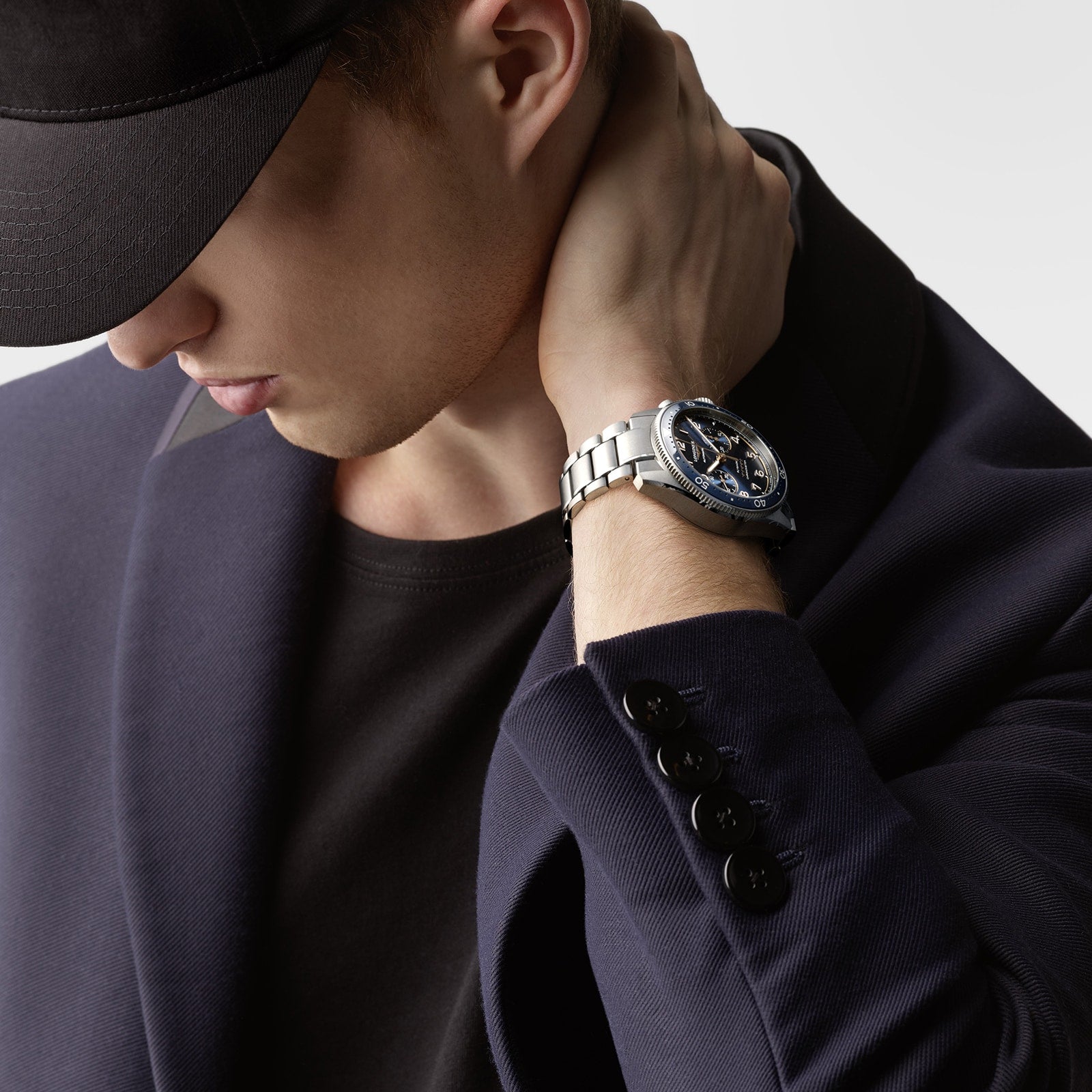 A person in a dark blazer and cap looks down, touching their neck, while wearing a Longines Spirit Flyback with a blue sunray dial and visible automatic calibre L791.